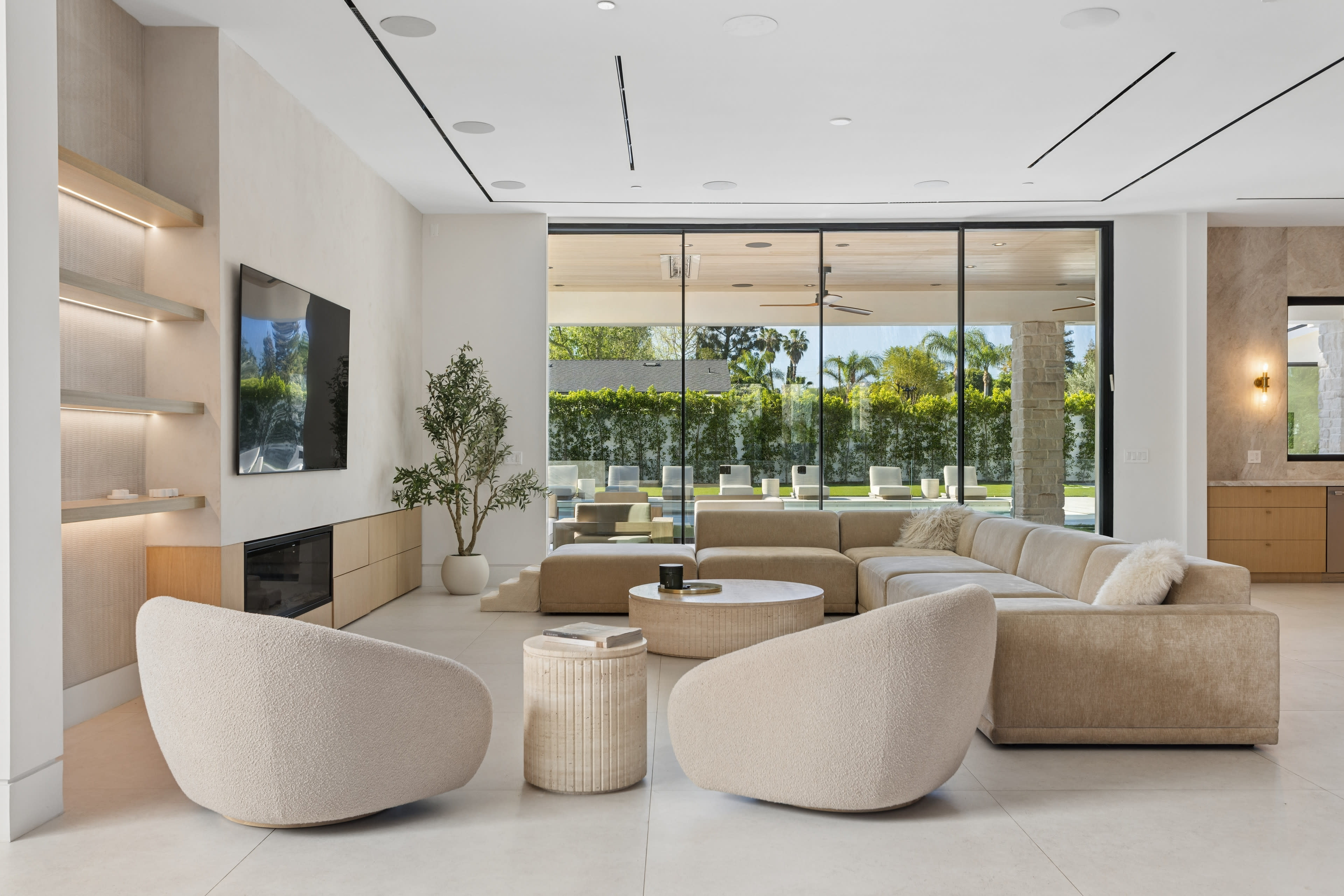 The image shows a modern living room with a large glass wall, a light-colored sectional sofa, two rounded chairs, and a coffee table, all surrounded by palm trees visible outside.