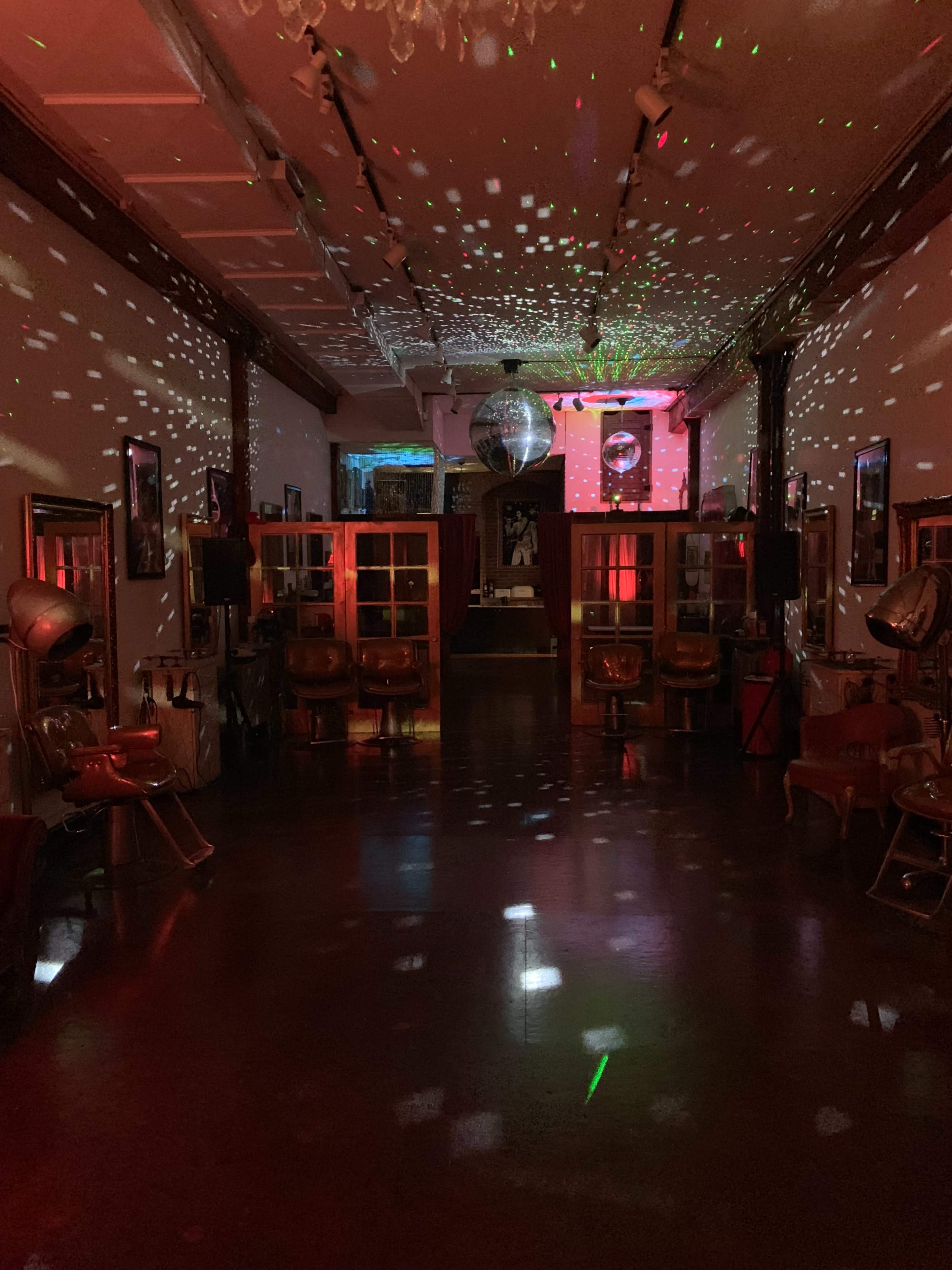 The image shows a dimly lit salon with red and green lights creating a disco effect, featuring vintage salon chairs and a large disco ball suspended from the ceiling.