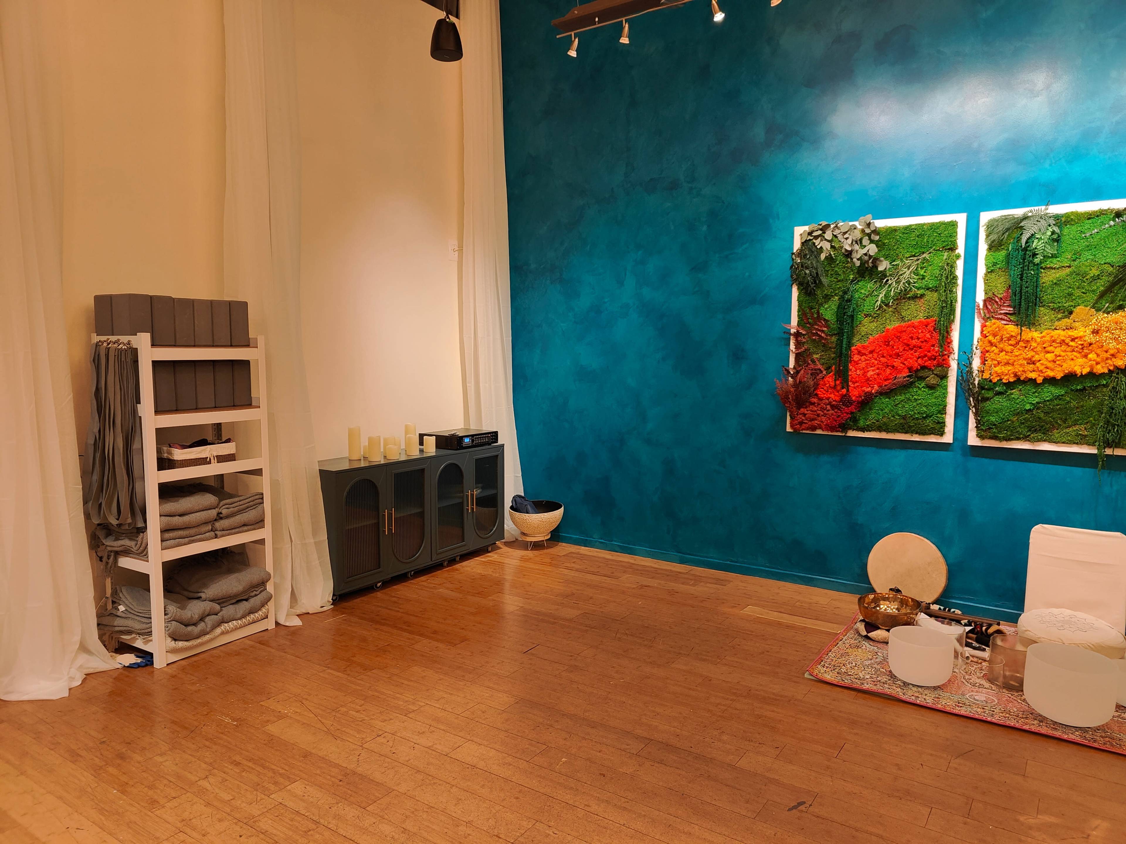 A serene indoor space features a turquoise wall, a shelf with neatly stacked towels, and two artistic wall hangings made of colorful natural materials.