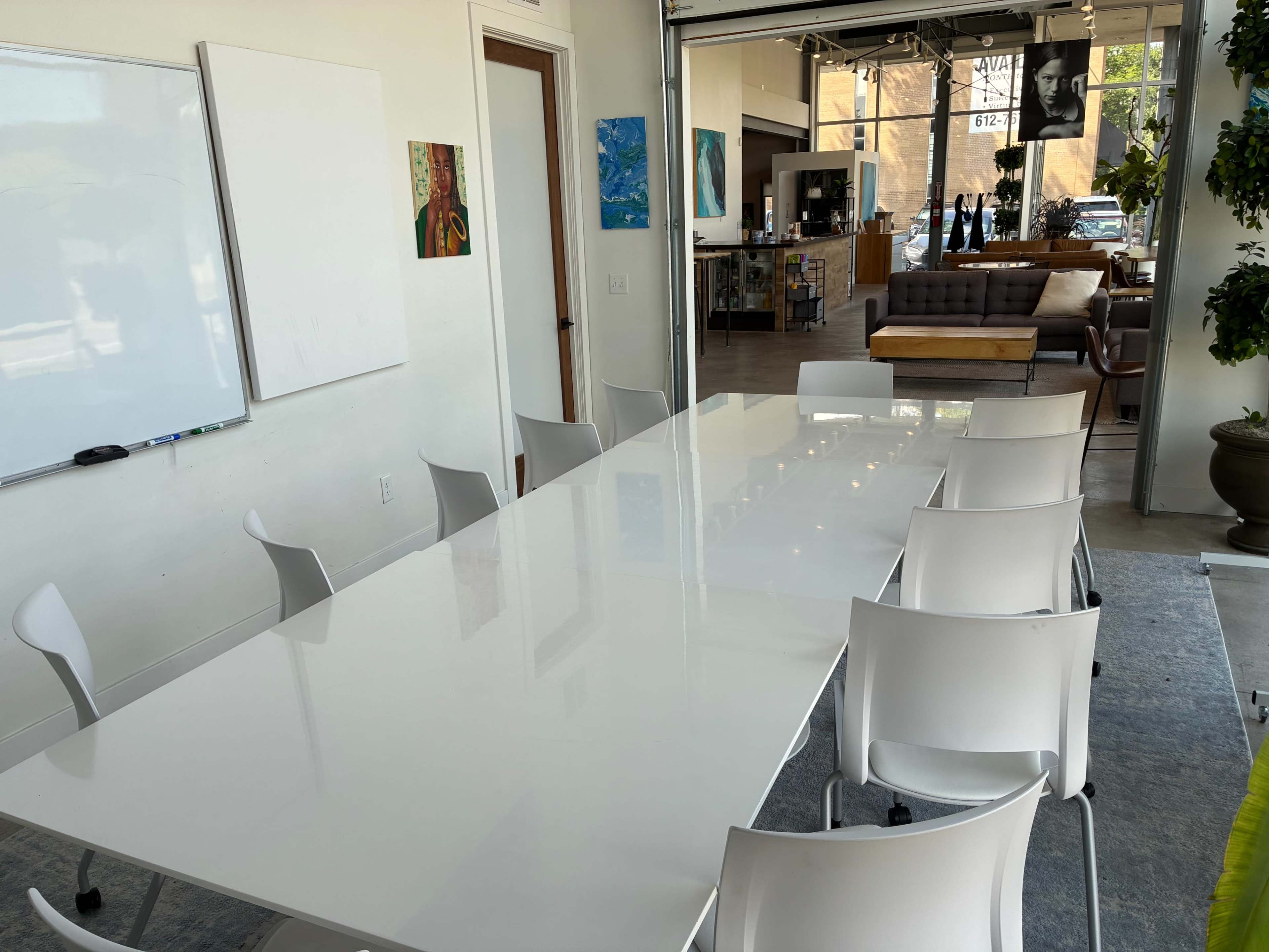The image shows a spacious meeting room with a long white table surrounded by white chairs, leading to a brightly lit area with a lounge and a café-style setting in the background.