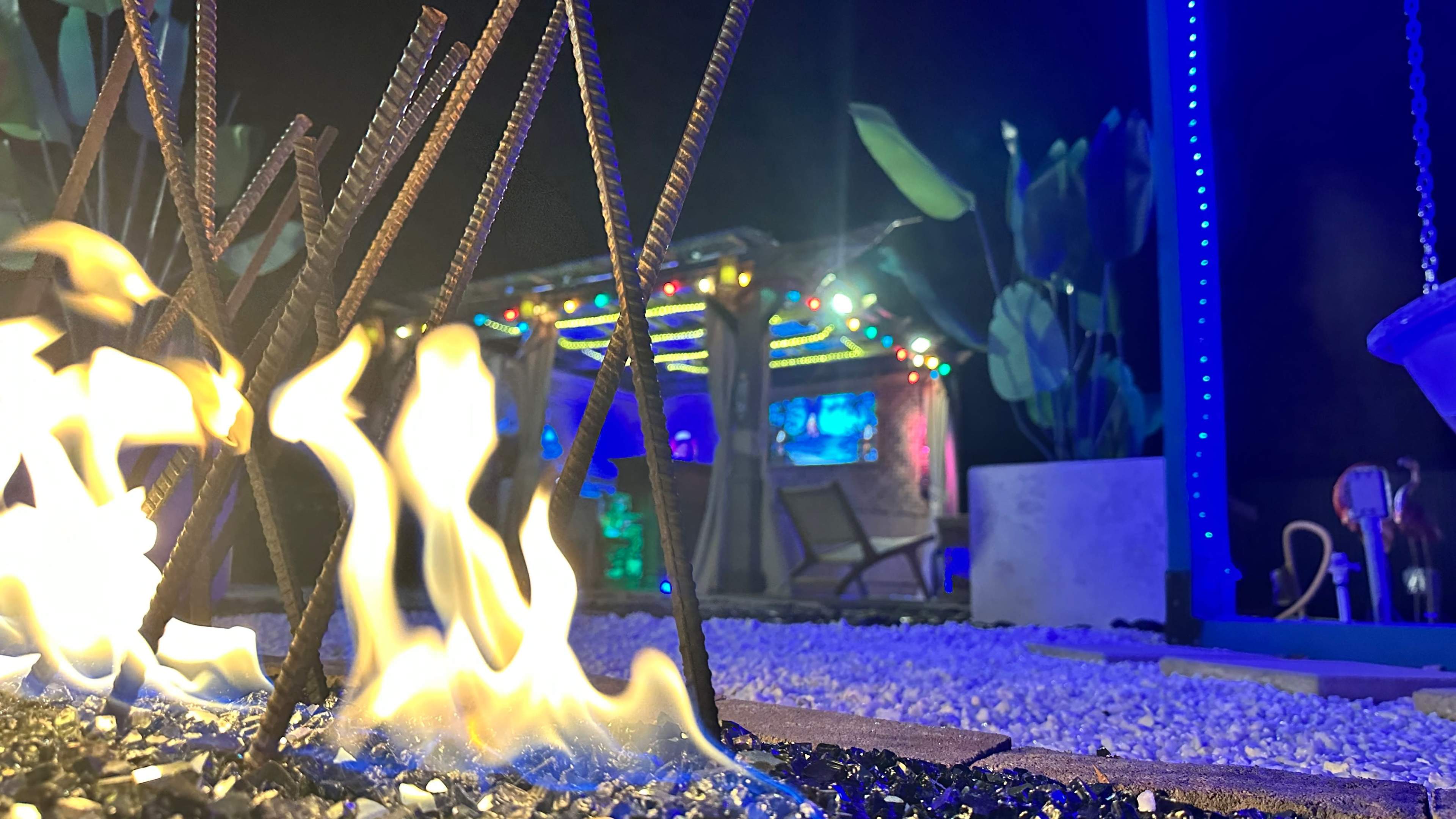 A brightly lit outdoor area features flames rising from a sculpture made of rebar, with a colorful bar structure and seating in the background.