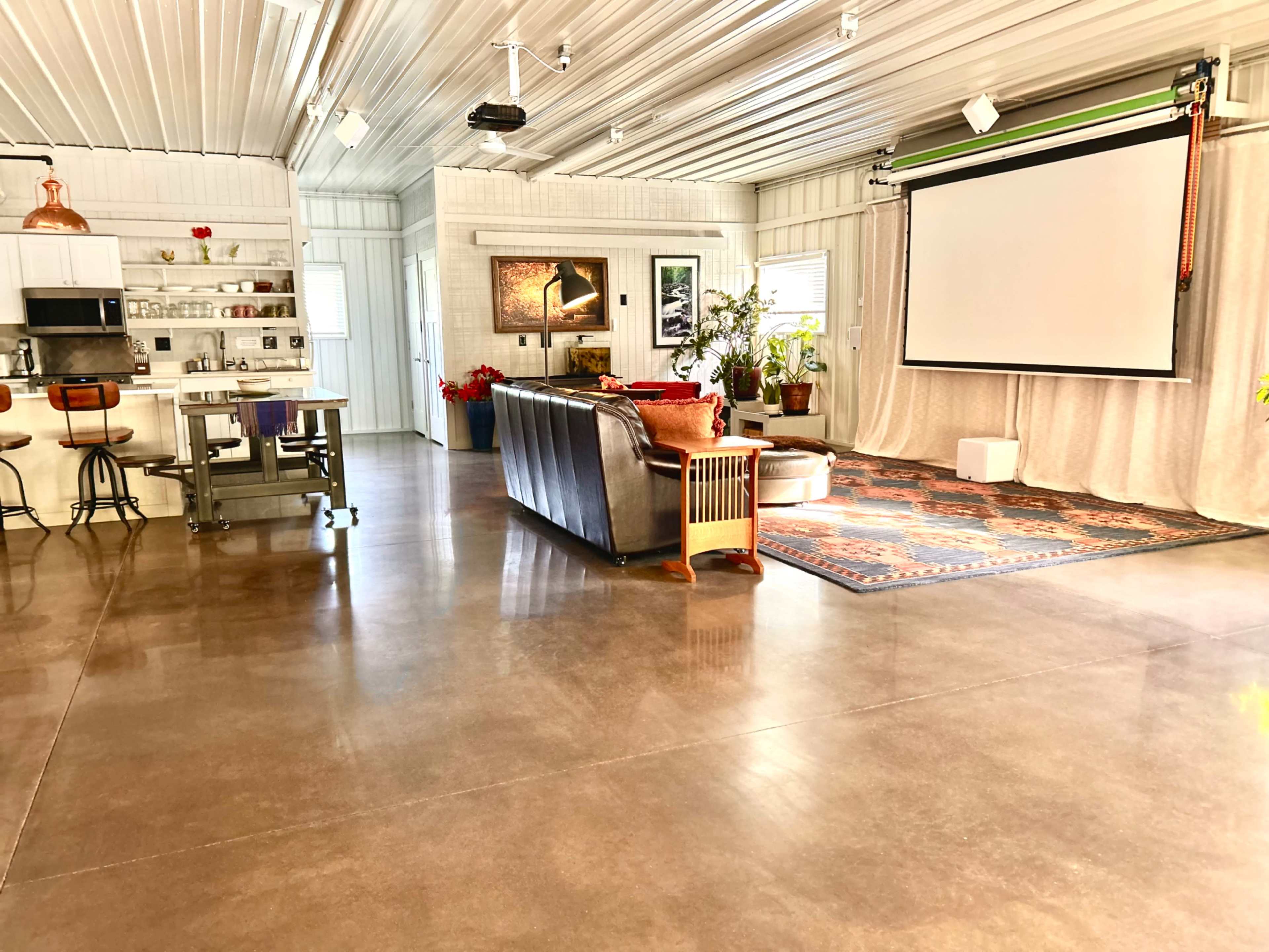 The image shows a spacious interior with a polished concrete floor, a seating area with a dark couch, a large screen on one wall, and a kitchen area featuring bar stools.