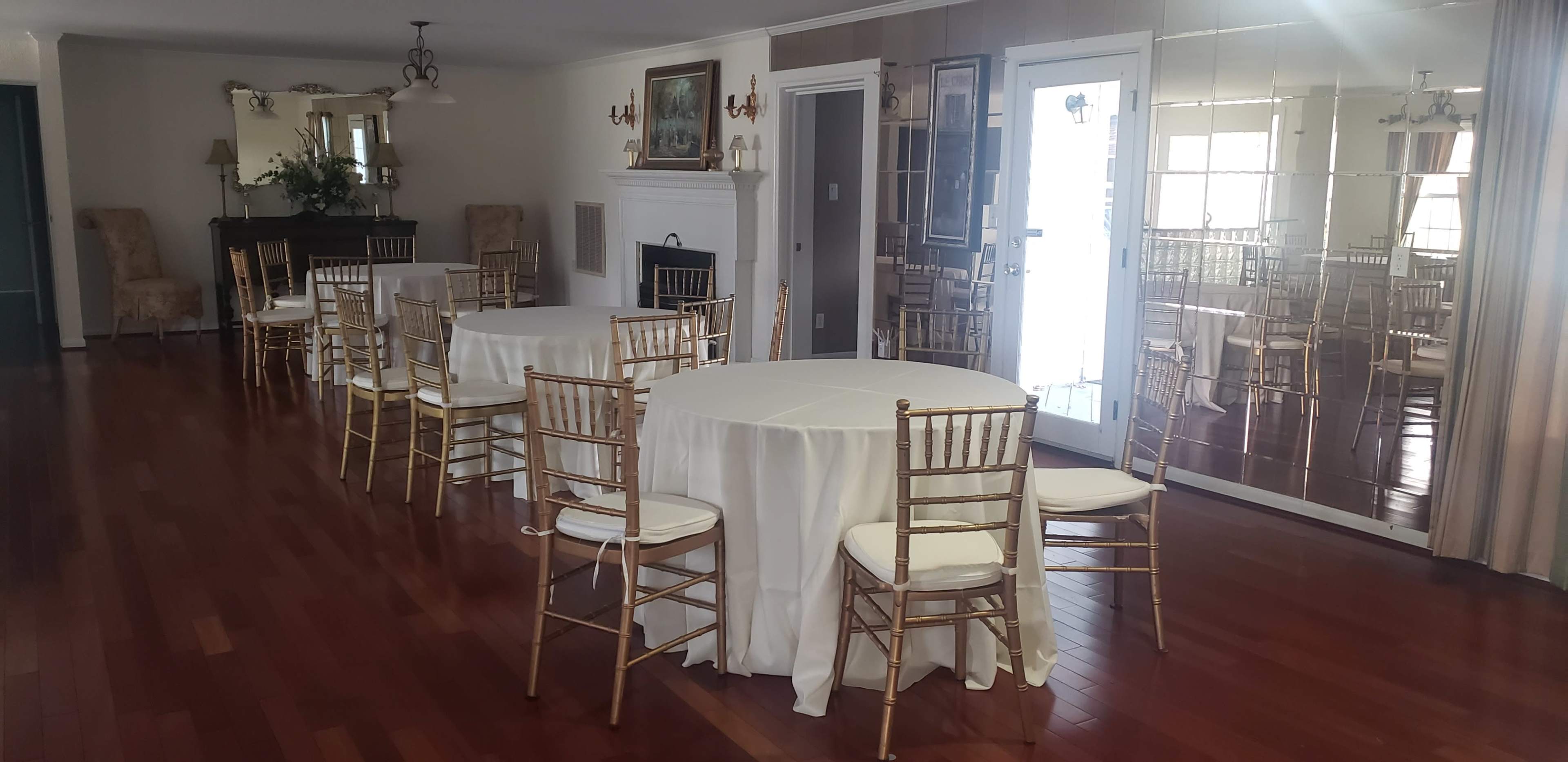 The room features several round tables with white tablecloths and gold chairs, arranged on a wooden floor, alongside a fireplace and large mirrors.