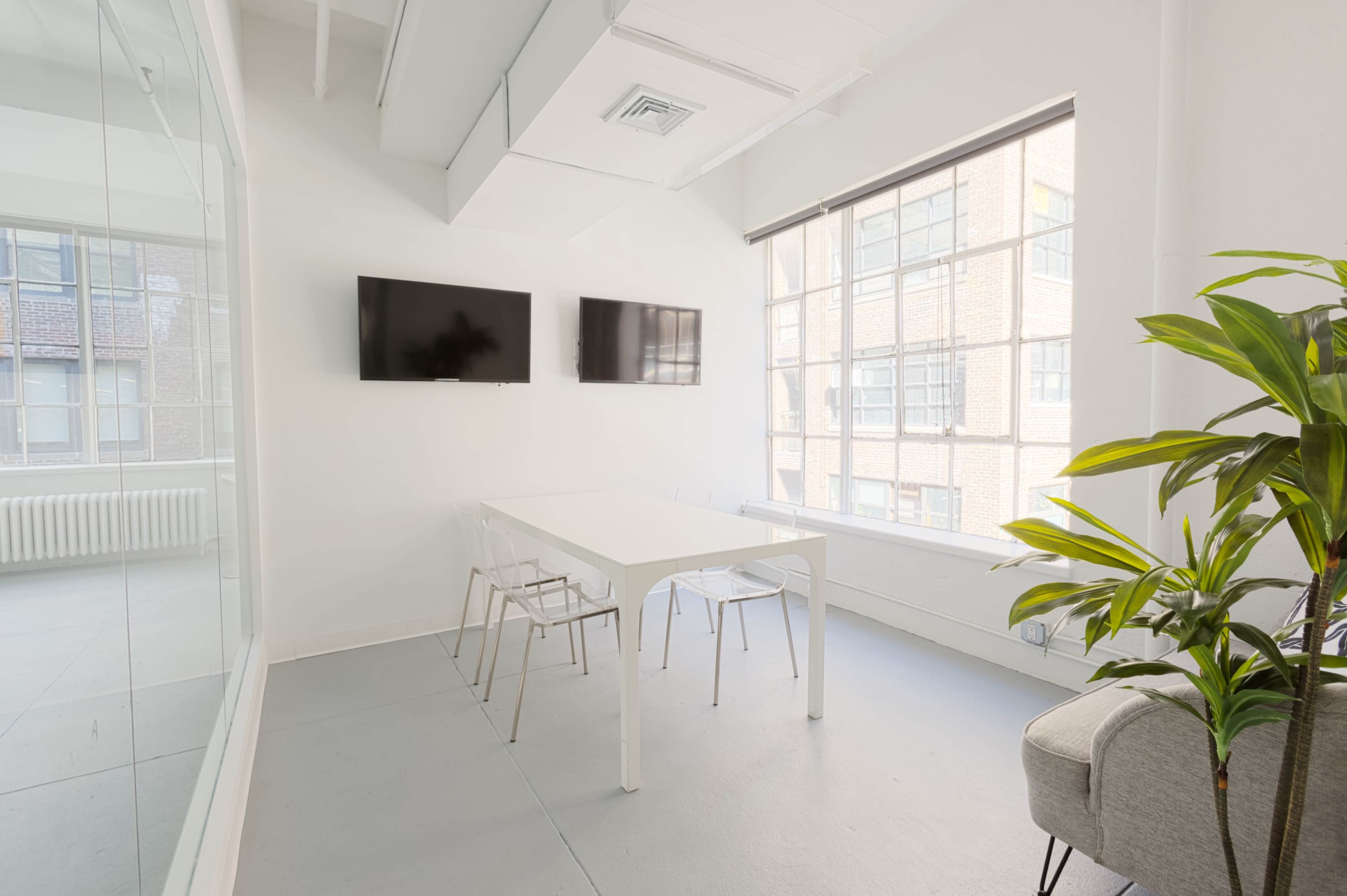 A bright meeting room with a white table, four transparent chairs, two wall-mounted televisions, and a large window with several panes.