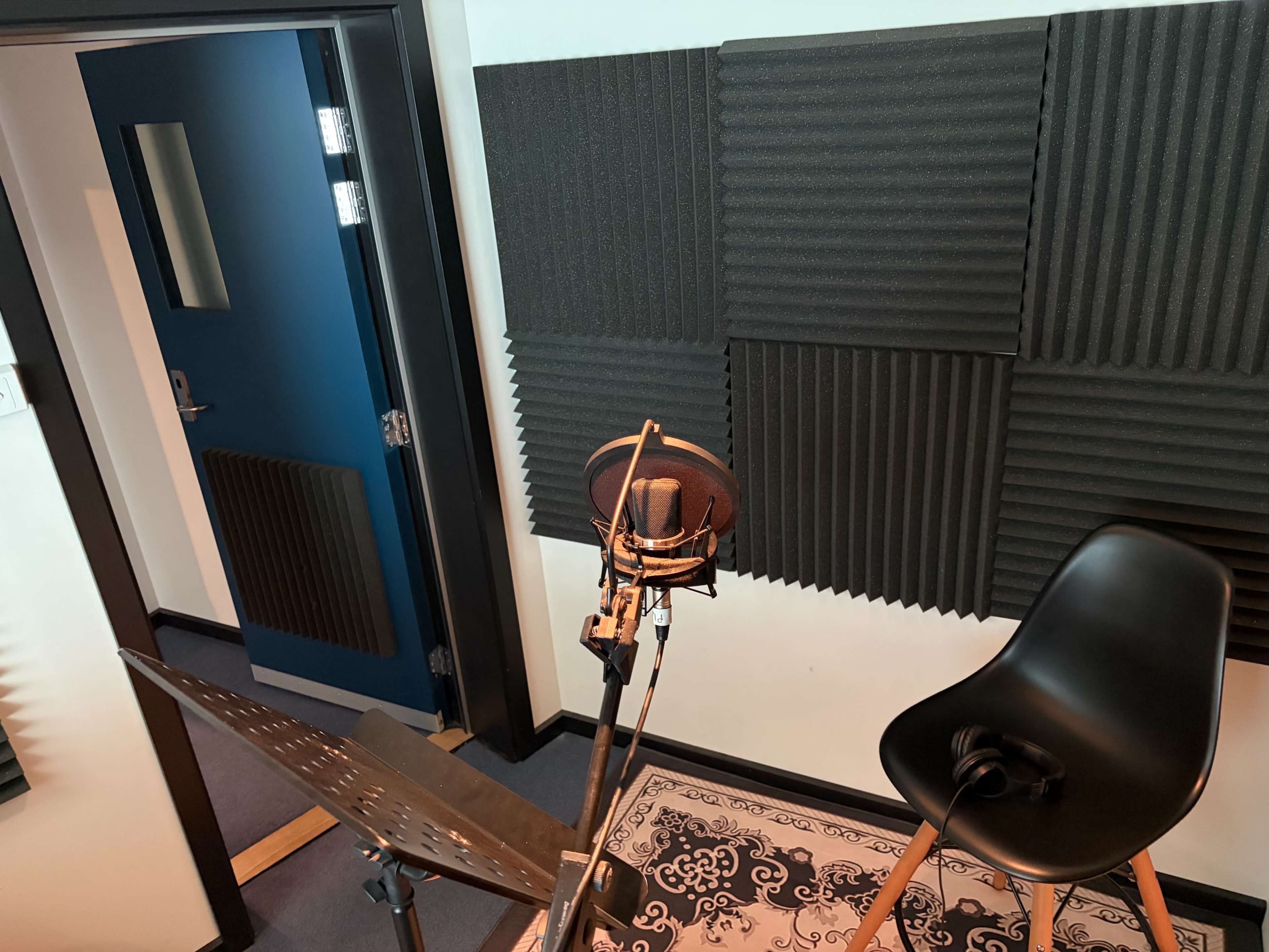 The image shows a recording booth with soundproof foam panels on the walls, a microphone on a stand, a chair, and a music stand.