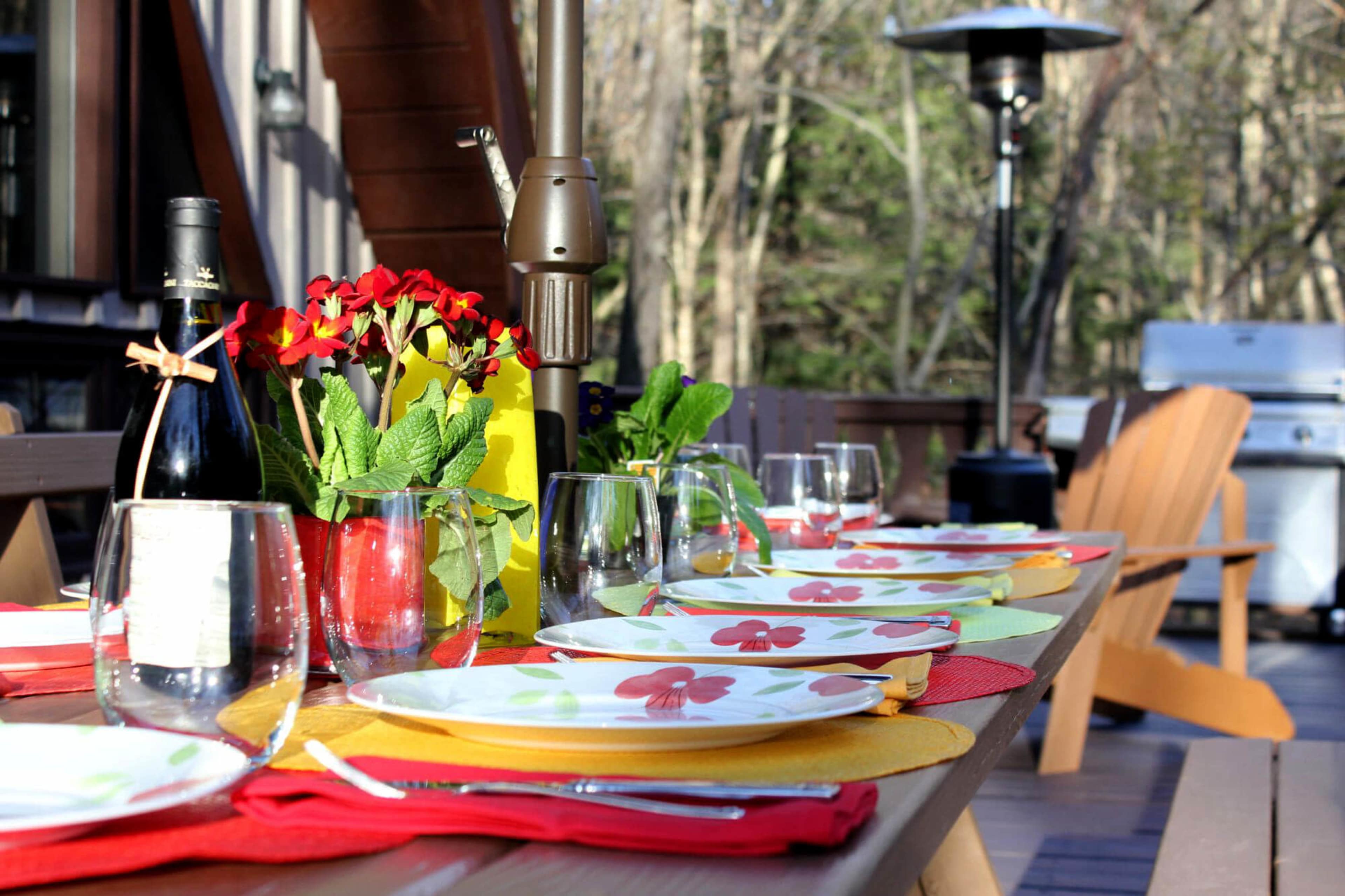 A long wooden table is set for a meal outdoors, featuring colorful plates, glasses, and a centerpiece of flowers, with a barbecue grill in the background.