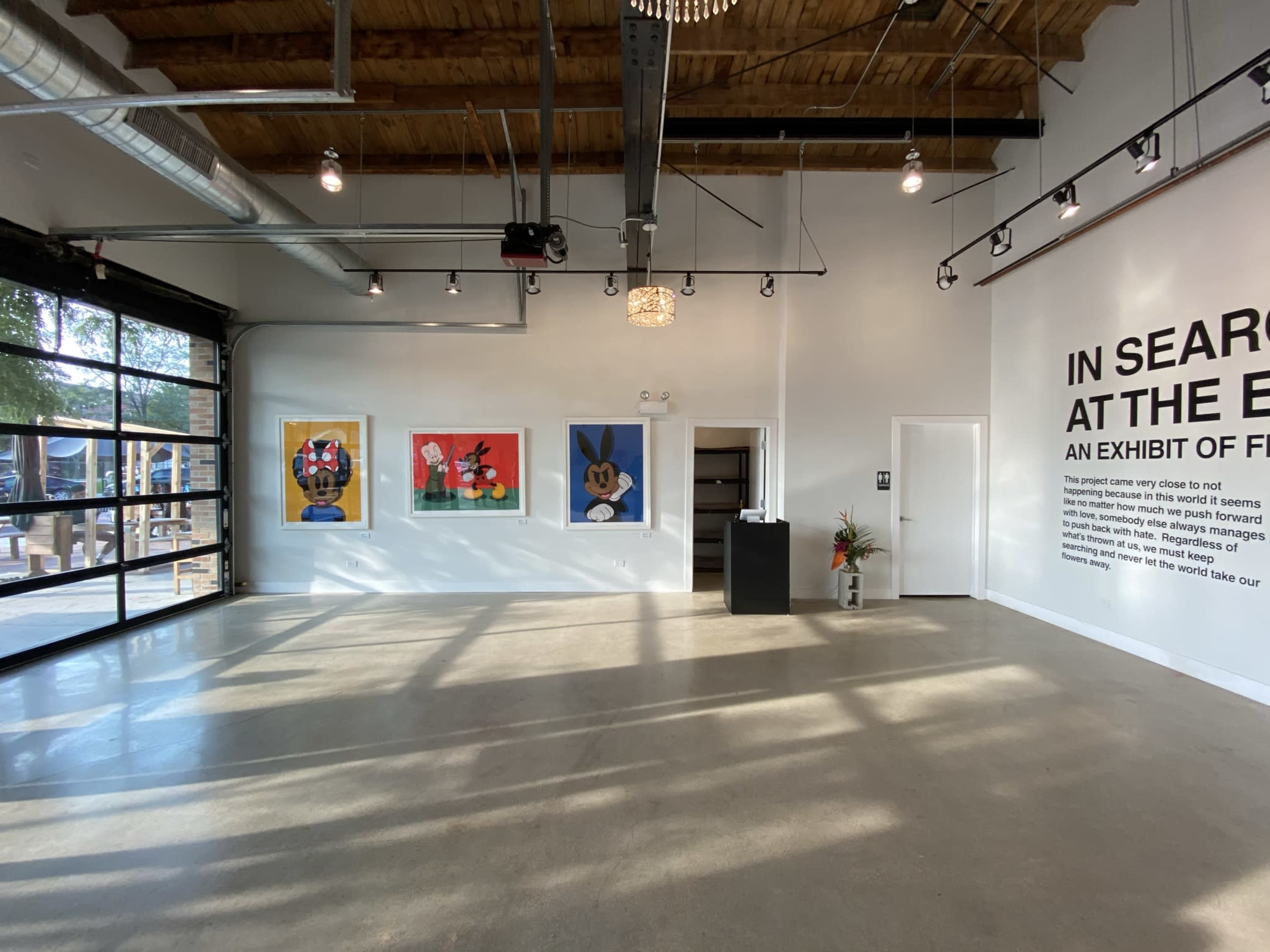 The image shows a spacious art exhibition space featuring three colorful paintings on the left wall and a large text display on the right wall.
