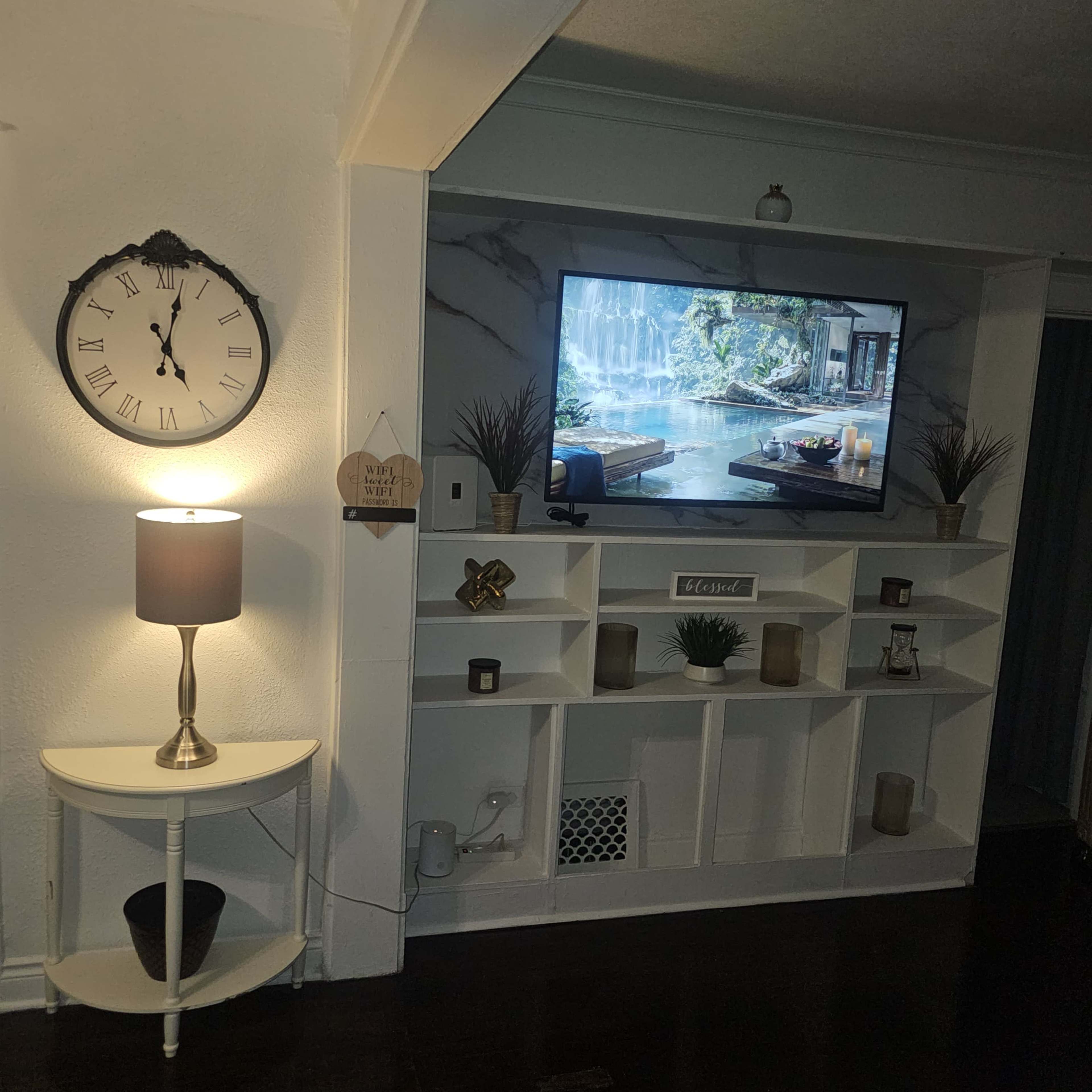A living room features a wall-mounted TV displaying a nature scene, a clock, a lamp on a small table, and a white shelving unit with decor items.