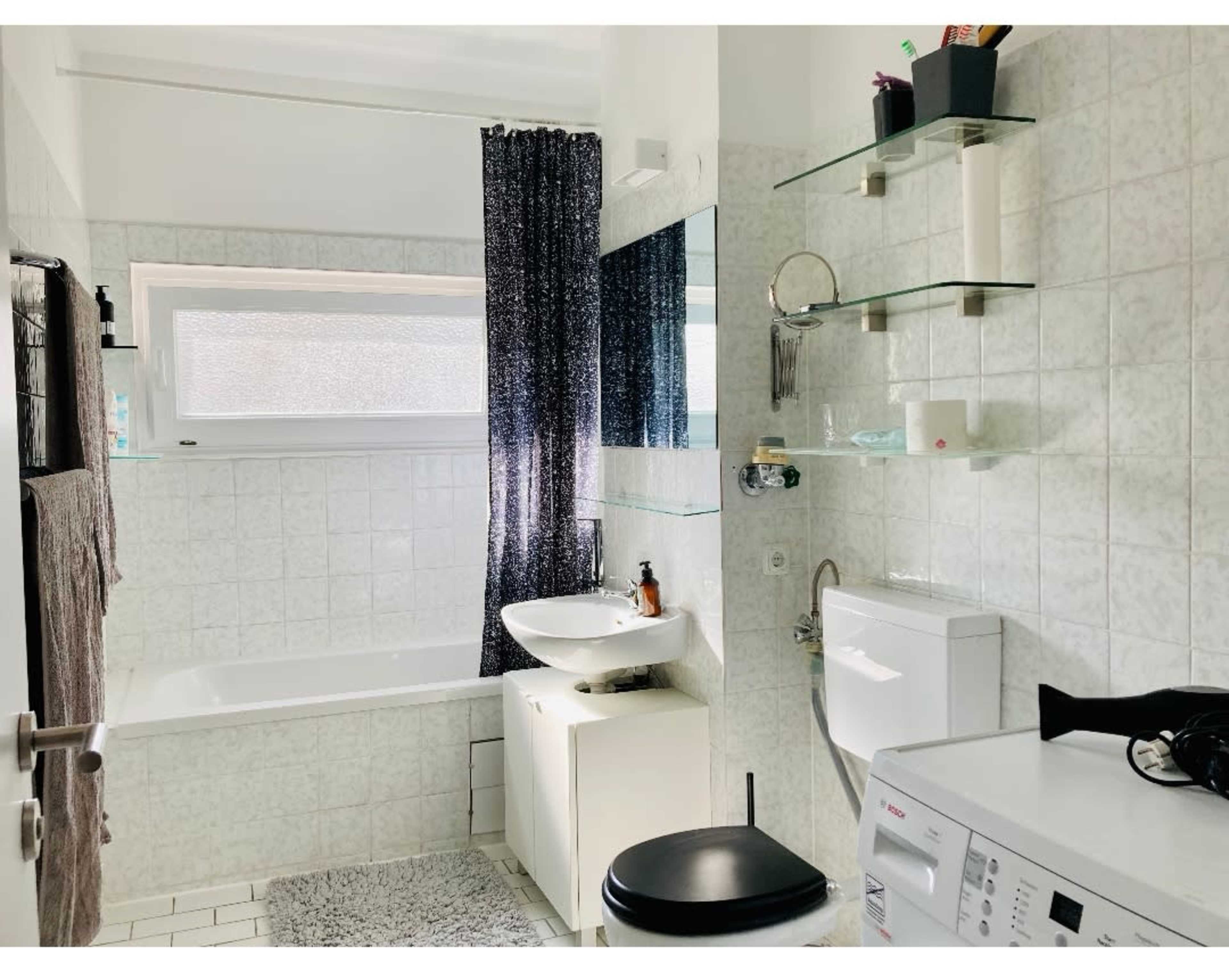 A bright bathroom featuring a bathtub, a sink with a mirror, a toilet, and a washing machine, all arranged against white tiled walls.