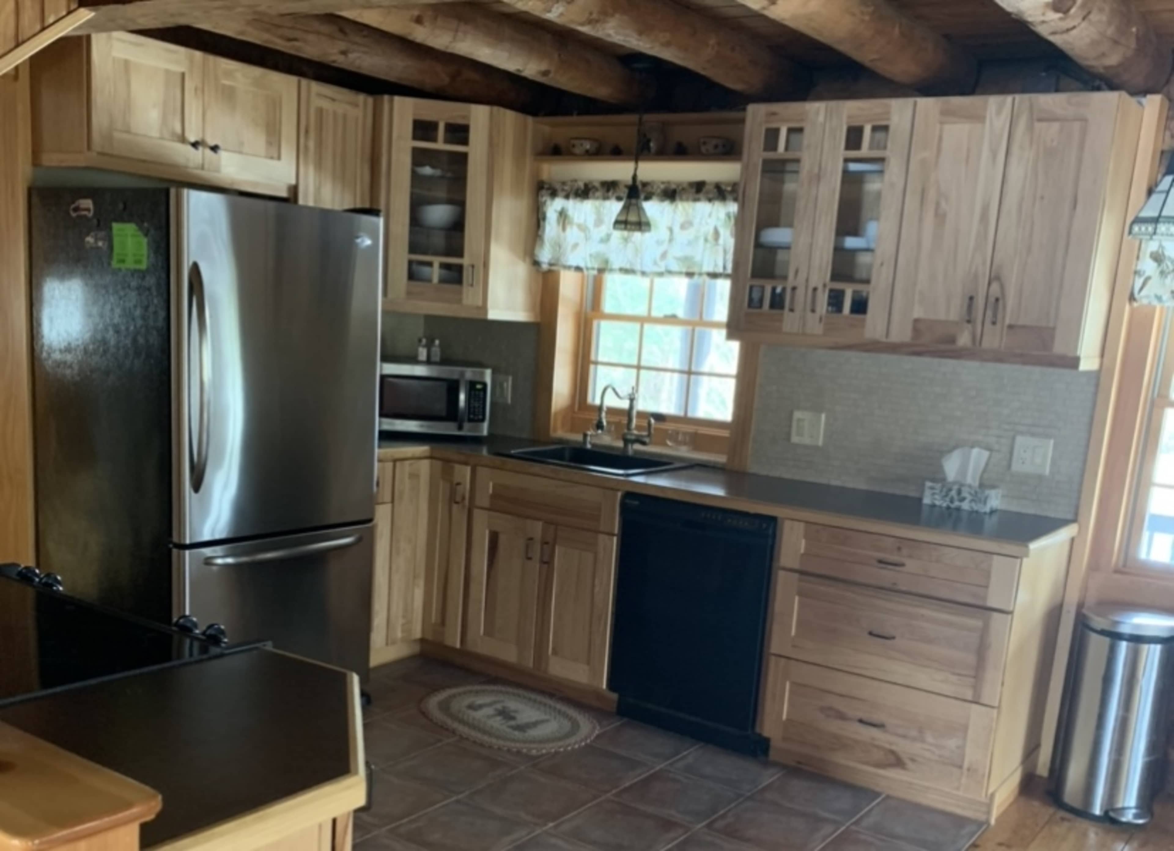 A wooden kitchen featuring stainless steel appliances, wooden cabinets, a dark countertop, and a window with a floral curtain.