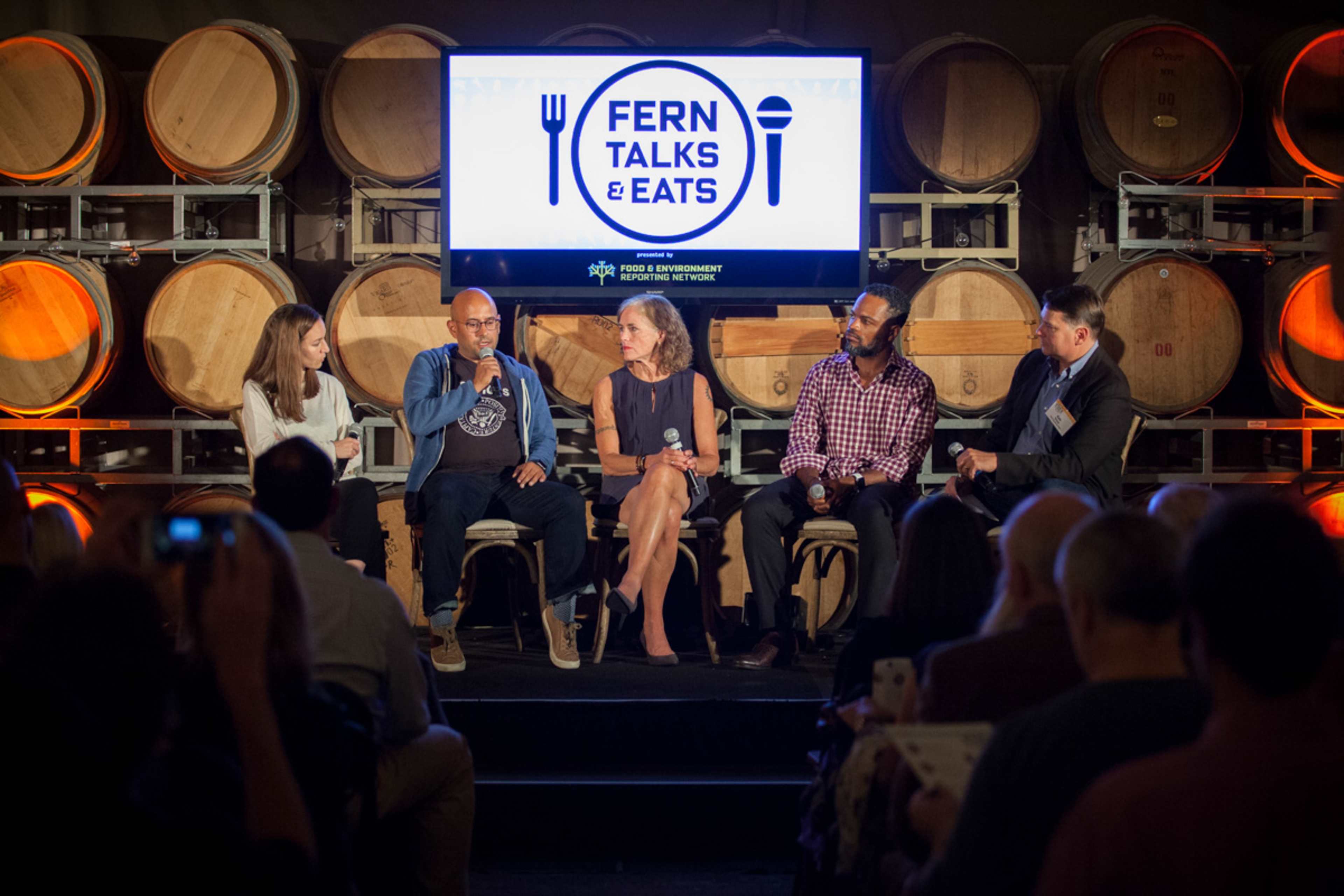 A panel discussion takes place on stage with five speakers seated in front of a screen, surrounded by wooden wine barrels.