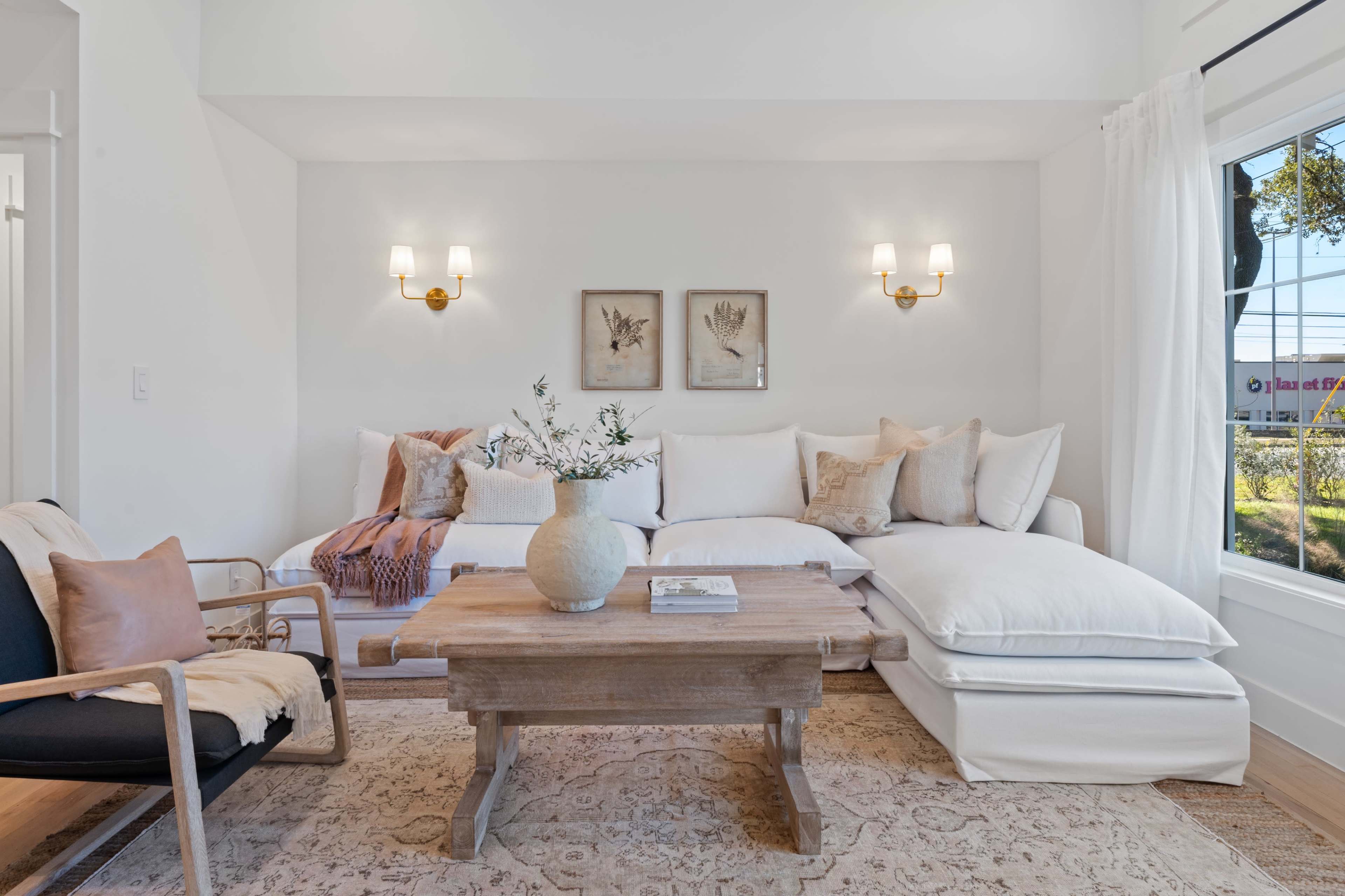 A bright, cozy living room features a sectional sofa with white cushions, a wooden coffee table, and decorative wall art.