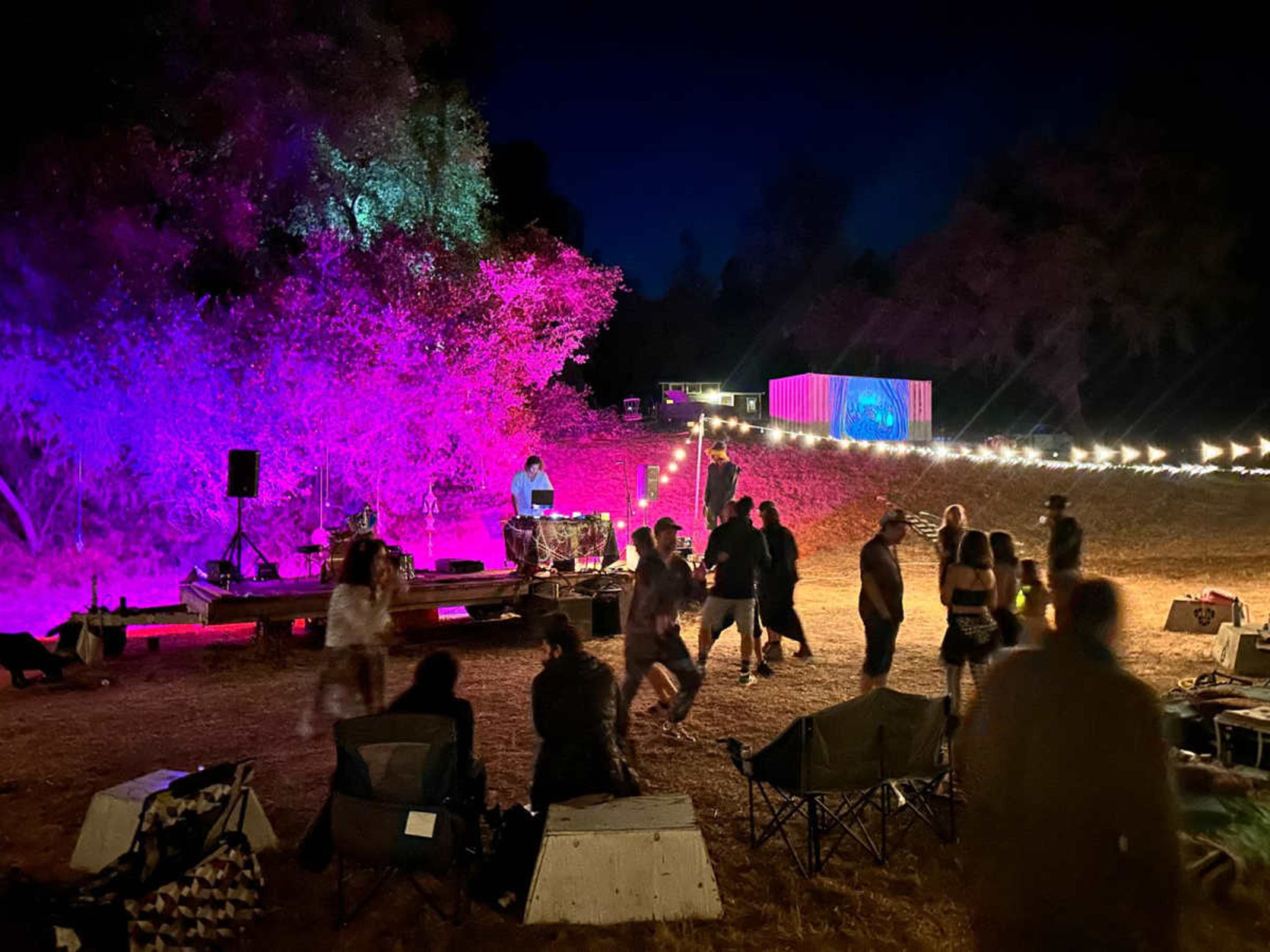 A DJ performs under colorful lighting while people dance and socialize in a gathering at night.