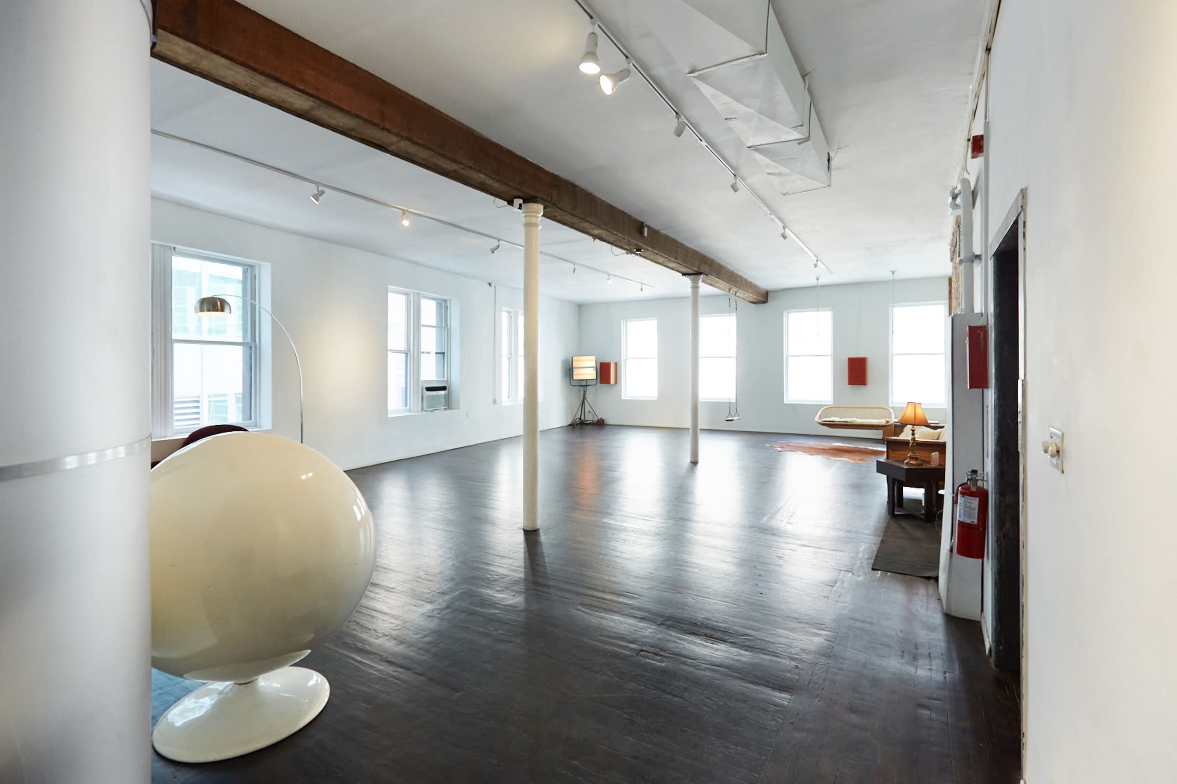 A spacious, empty room with wooden floors, large windows, and minimal furniture including a lamp and a table.