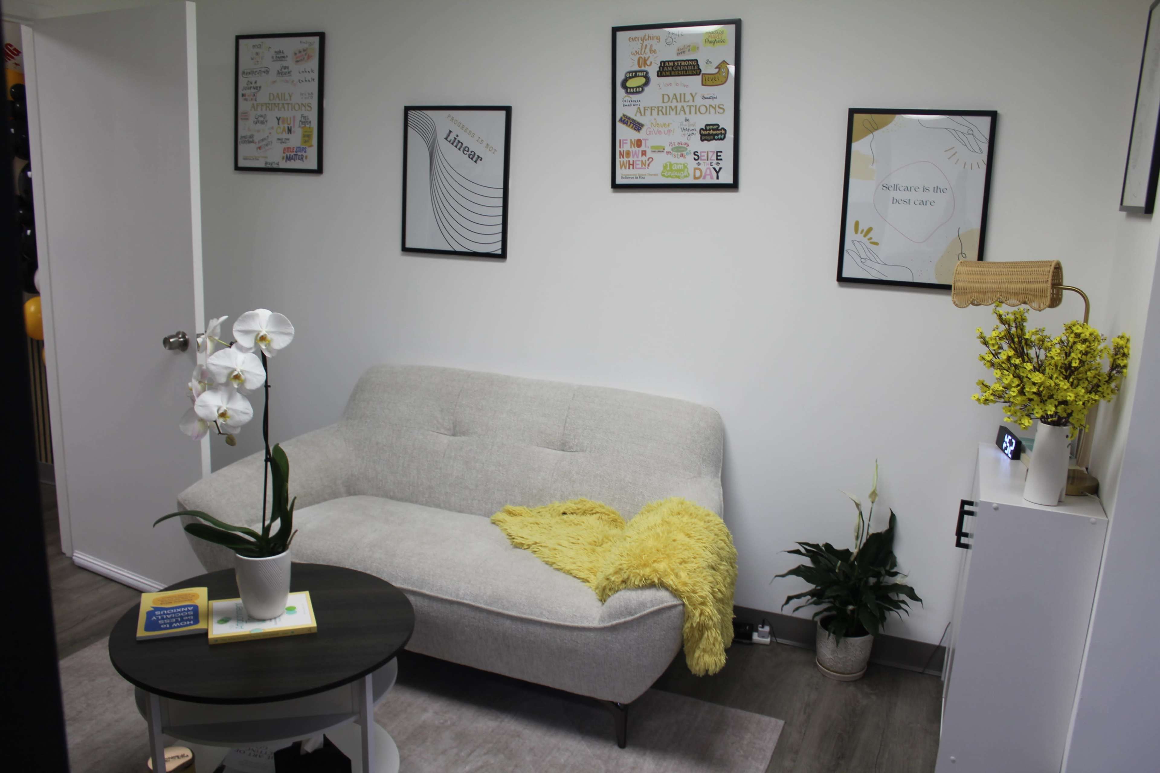 The image shows a modern, minimalist room featuring a light gray sofa with a yellow throw blanket, a round table, decorative plants, and framed motivational posters on the wall.