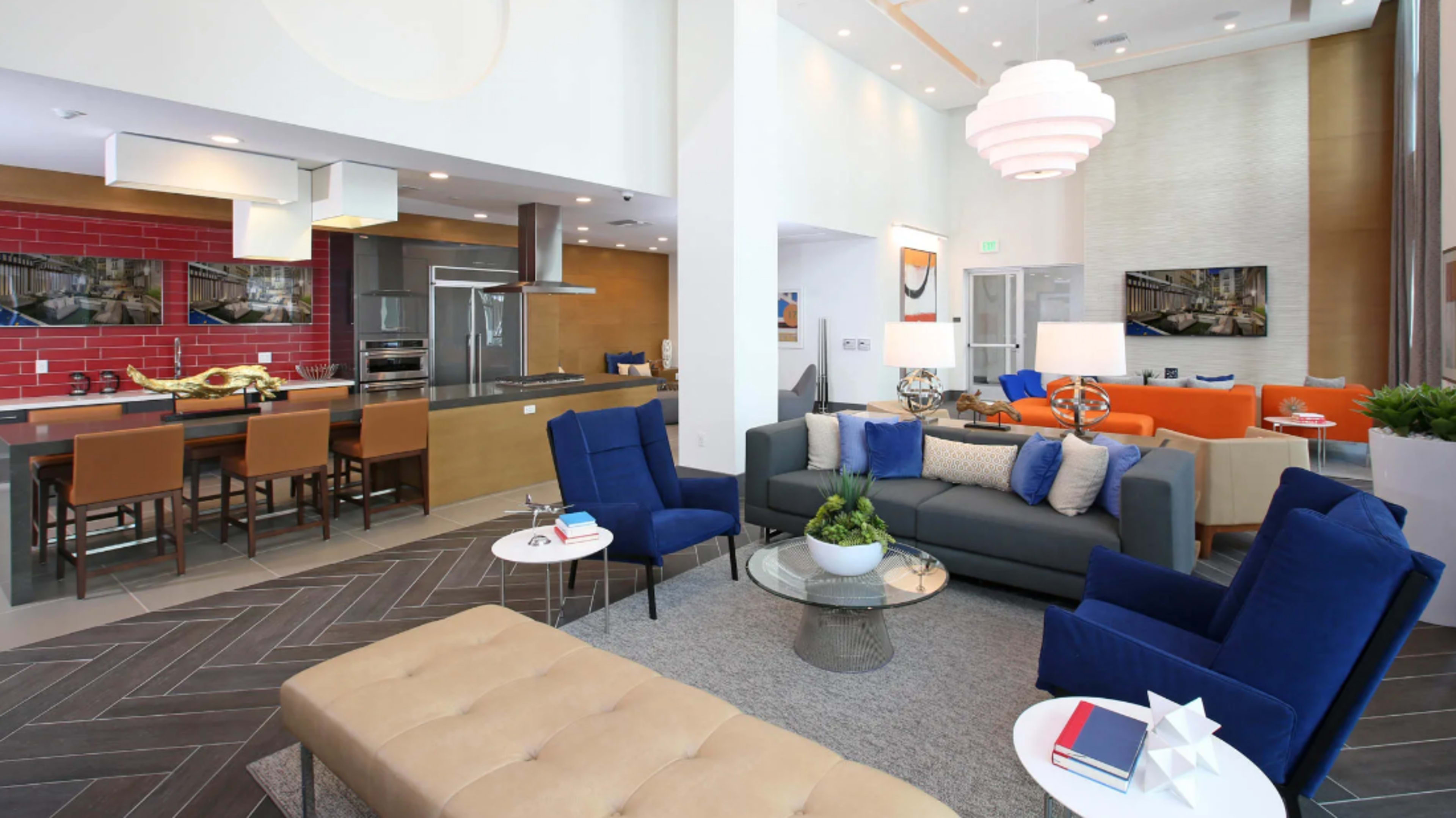 A modern living space featuring a mix of seating areas with colorful furniture, a kitchen with a red backsplash, and large windows allowing natural light.