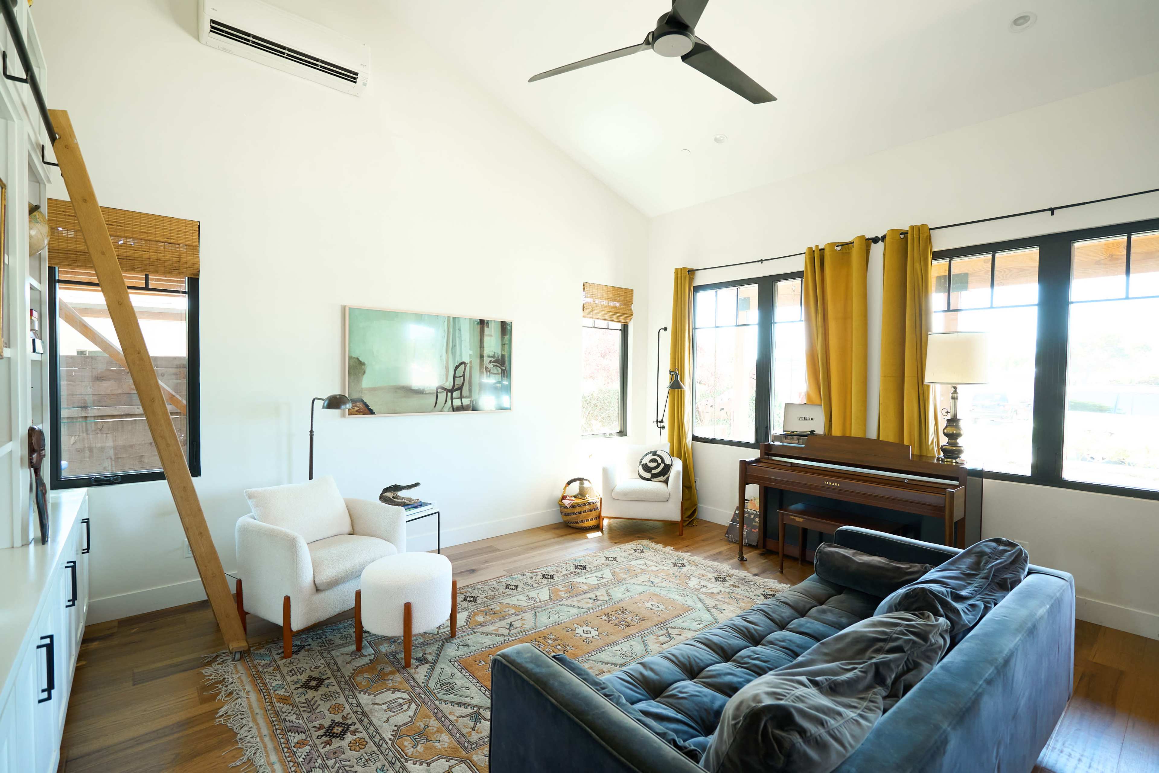 The living room features a large window, a piano, a blue sofa, and two white armchairs, with wooden flooring and a patterned rug.