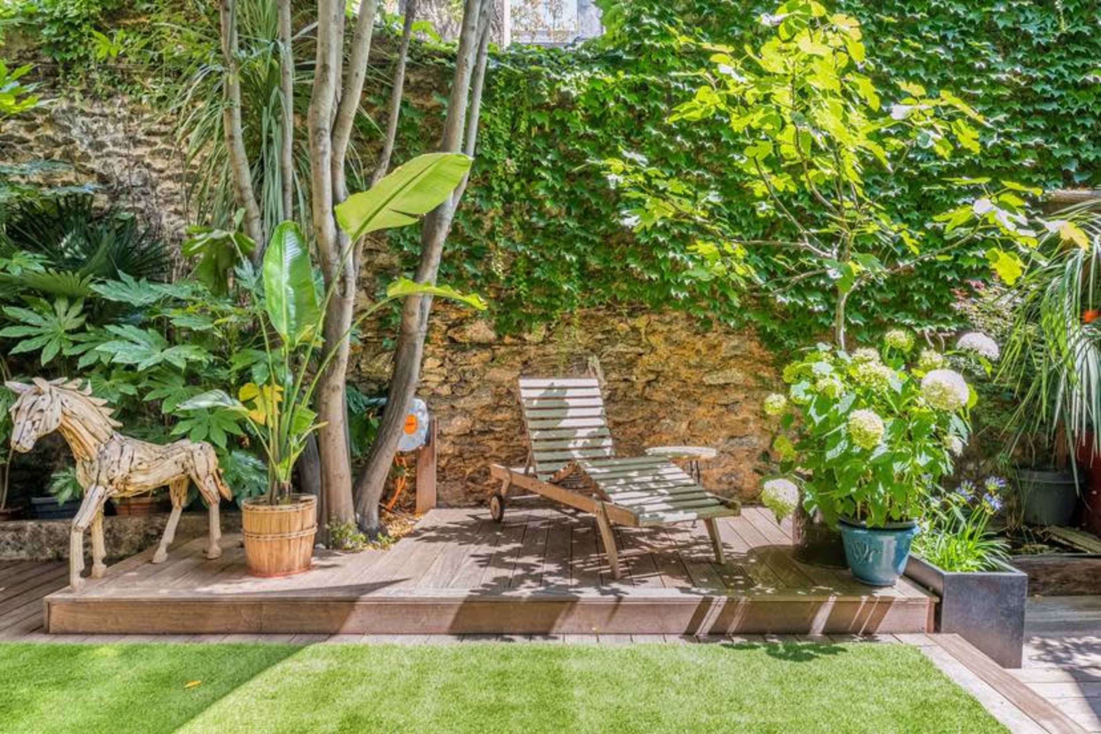 A wooden deck with a reclining lounge chair is surrounded by lush greenery and potted plants, including a horse sculpture.