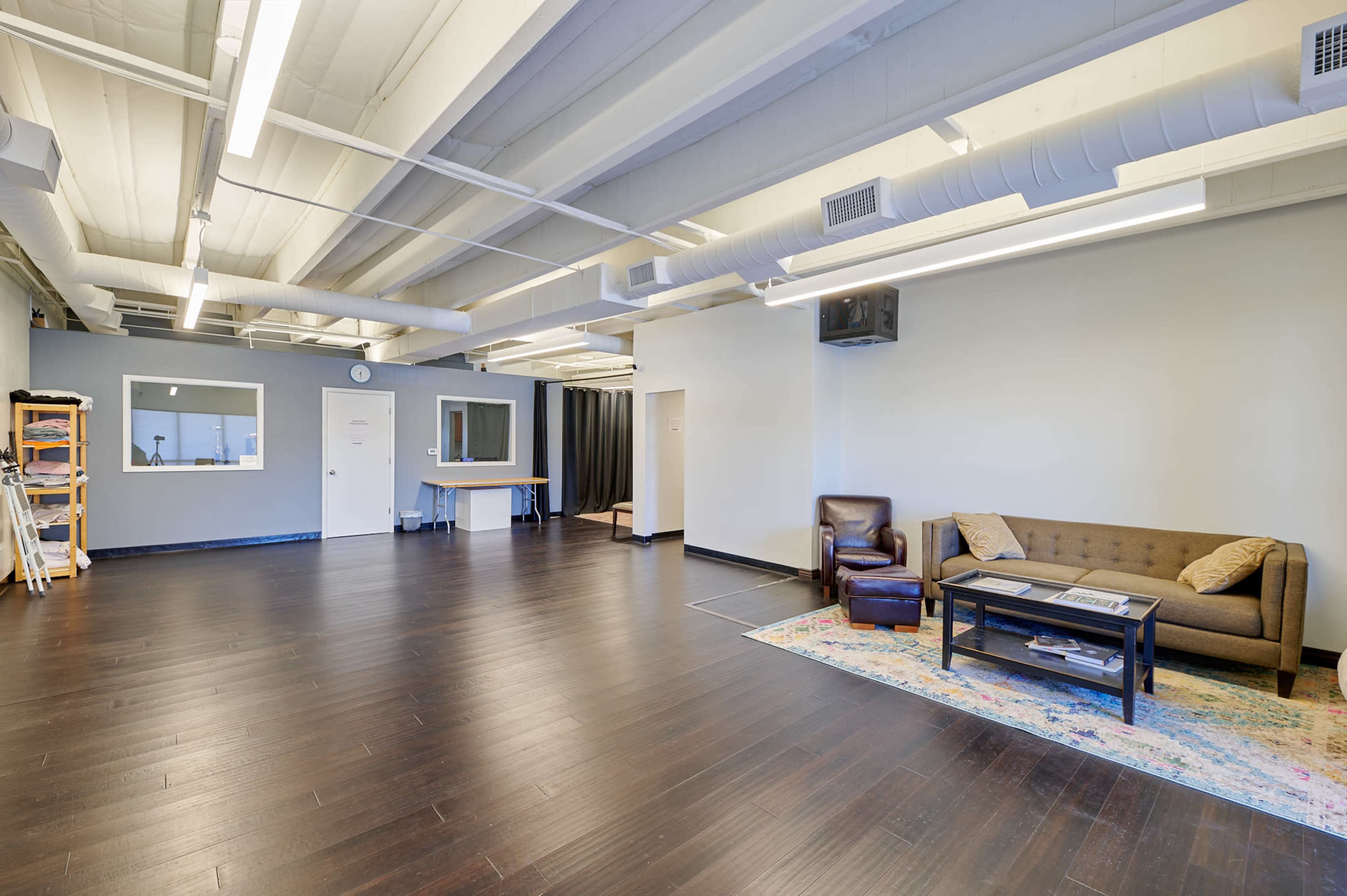 A spacious, unfurnished room with dark wooden floors, a couch, a coffee table, and a small kitchenette area.