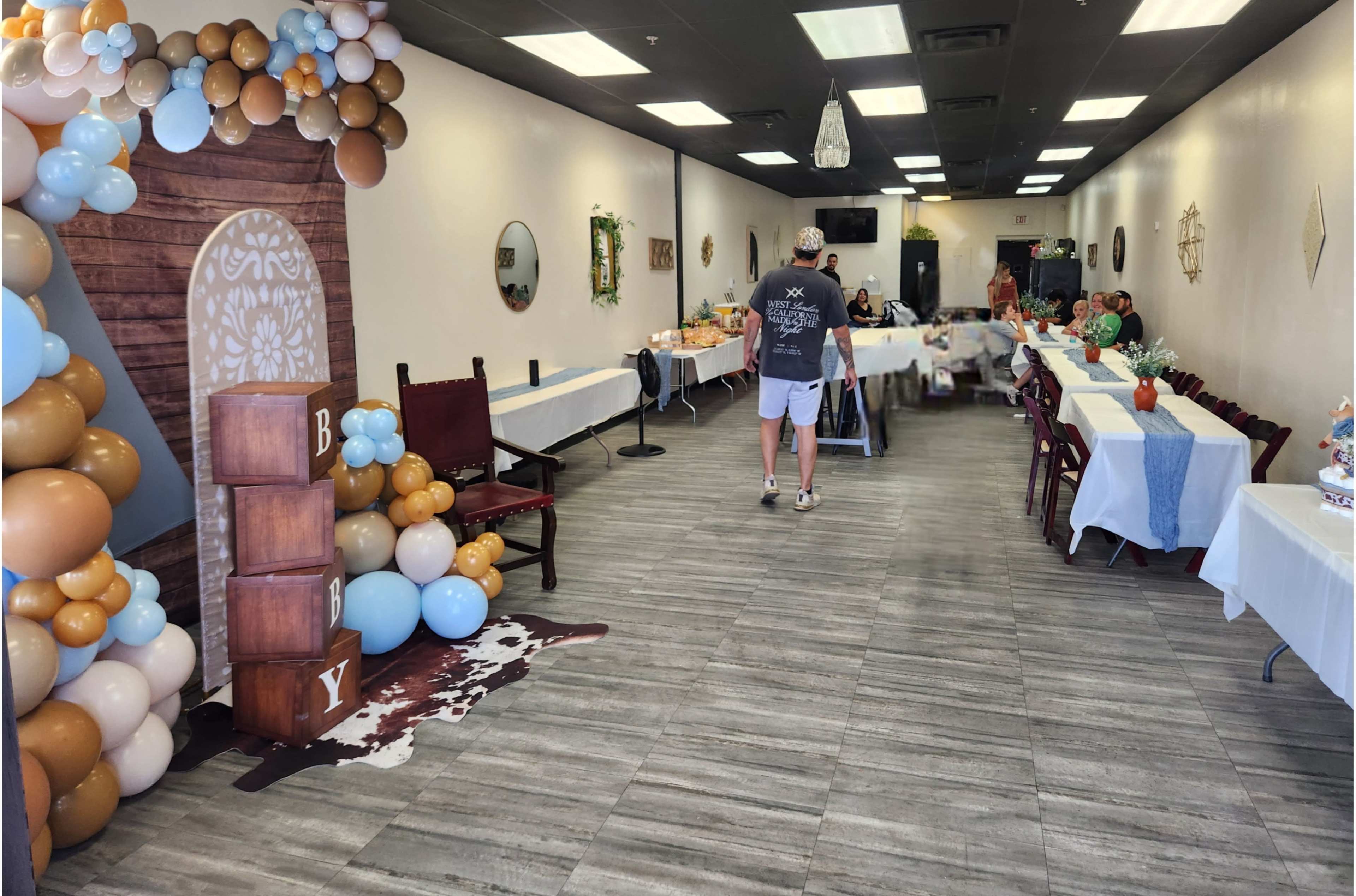 The image shows a decorated indoor event space with white tables, a balloon arch, and a person walking towards the back of the room.
