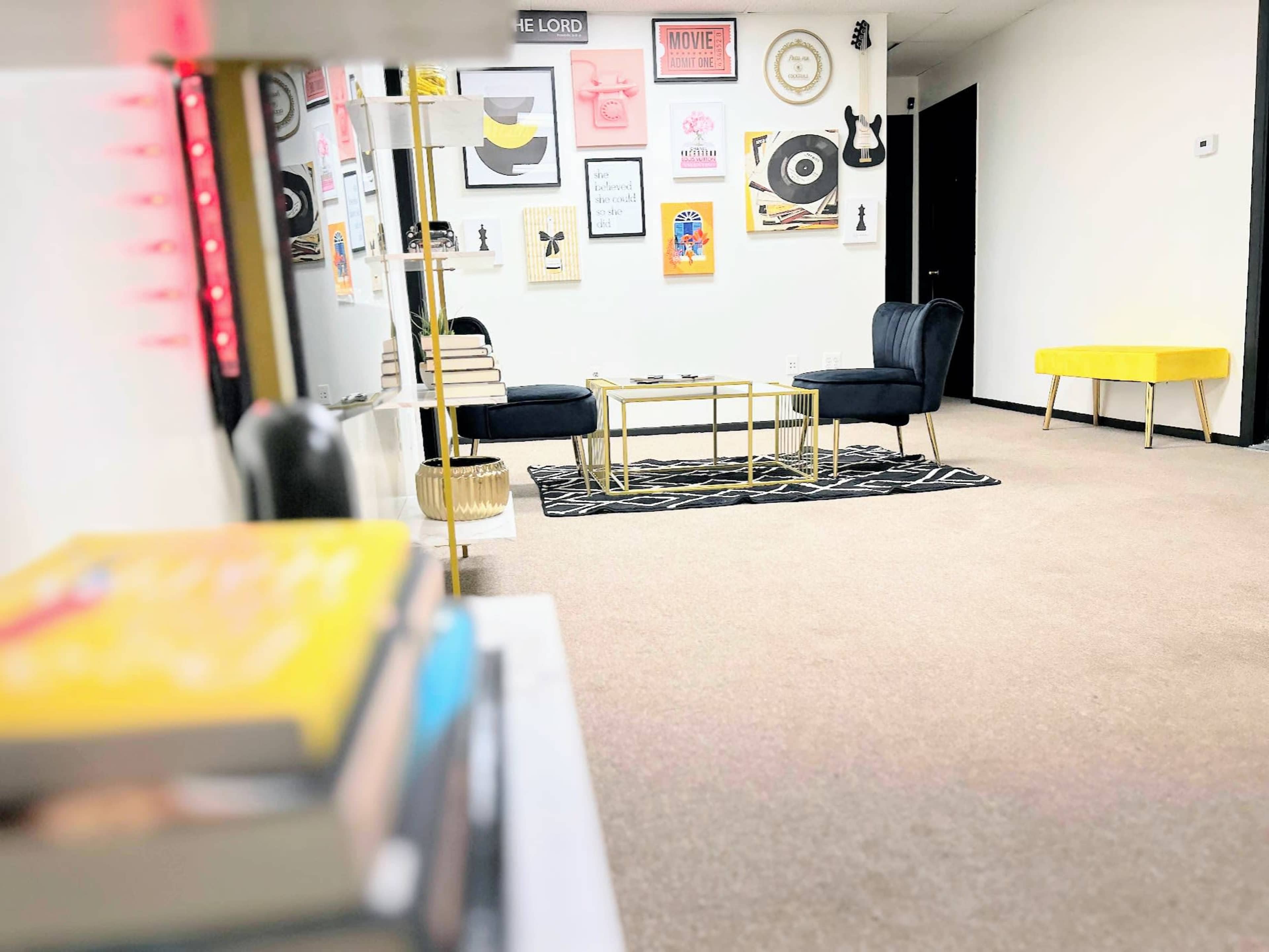A modern, minimalist room with a yellow accent table, a black chair, framed artwork on the walls, and a patterned rug on the floor.