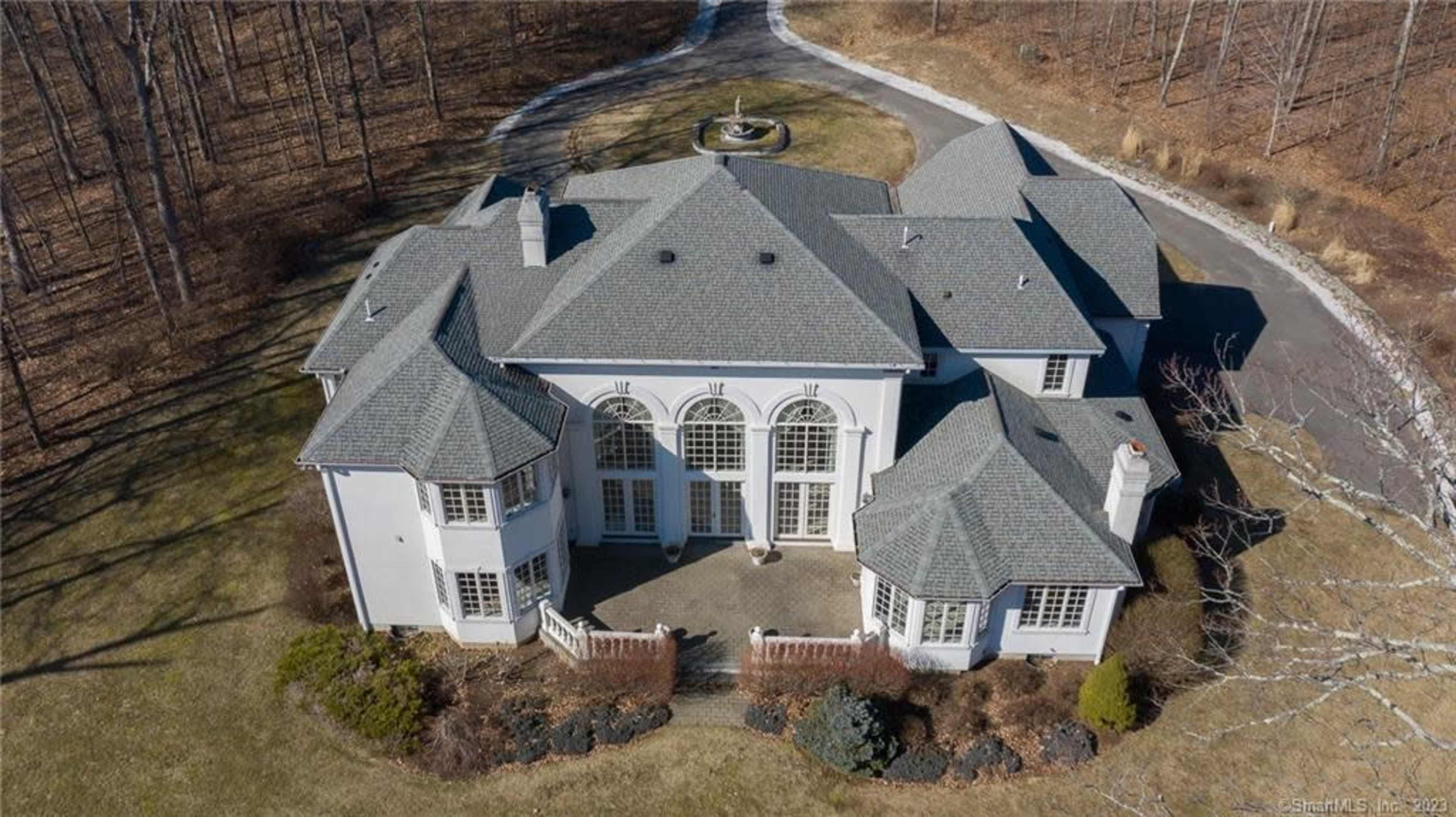 The image shows an aerial view of a large, white residential home surrounded by landscaped grounds and trees.