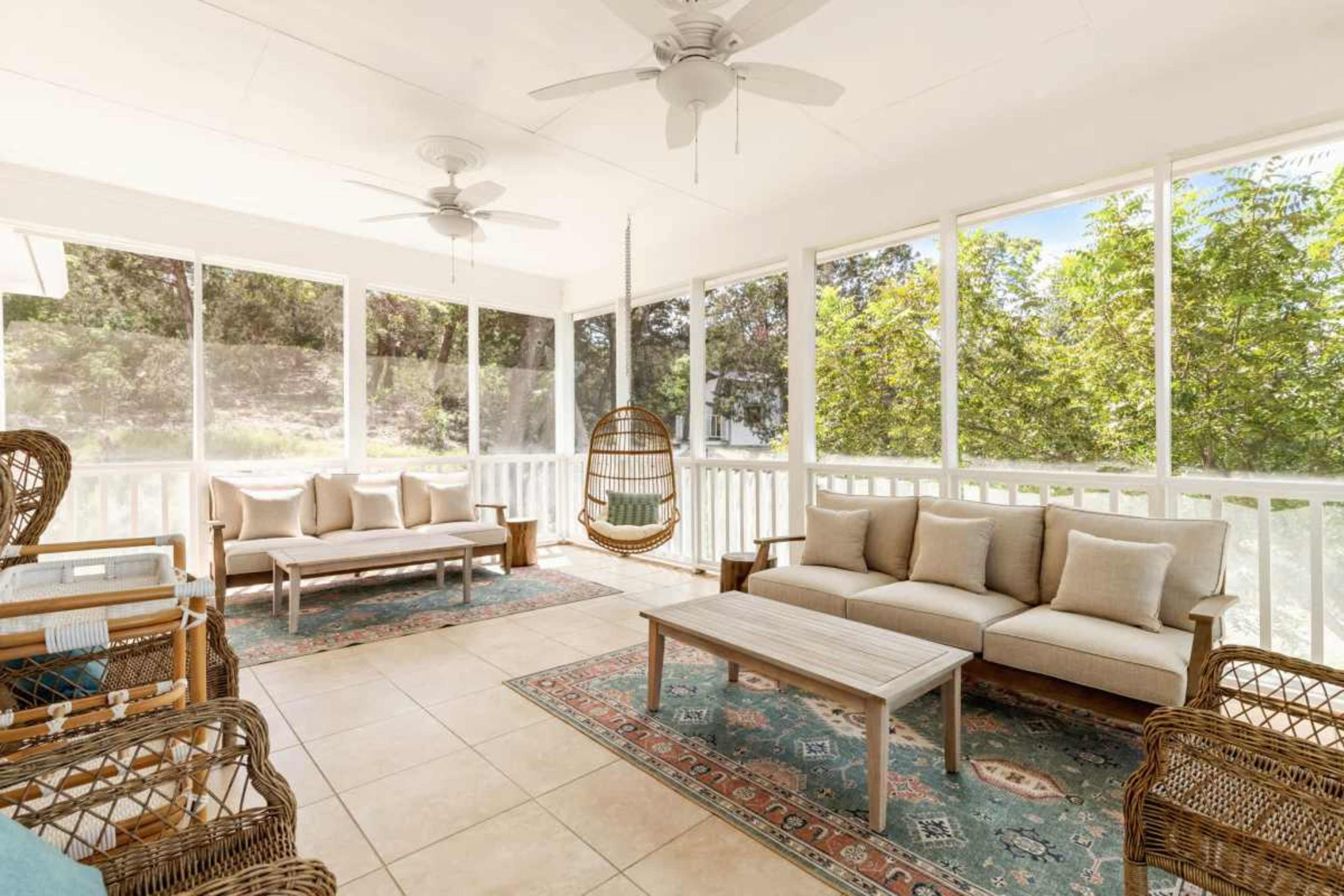 The image shows a screened-in porch with a seating area featuring wicker furniture, a hanging swing chair, and a coffee table on a patterned rug.