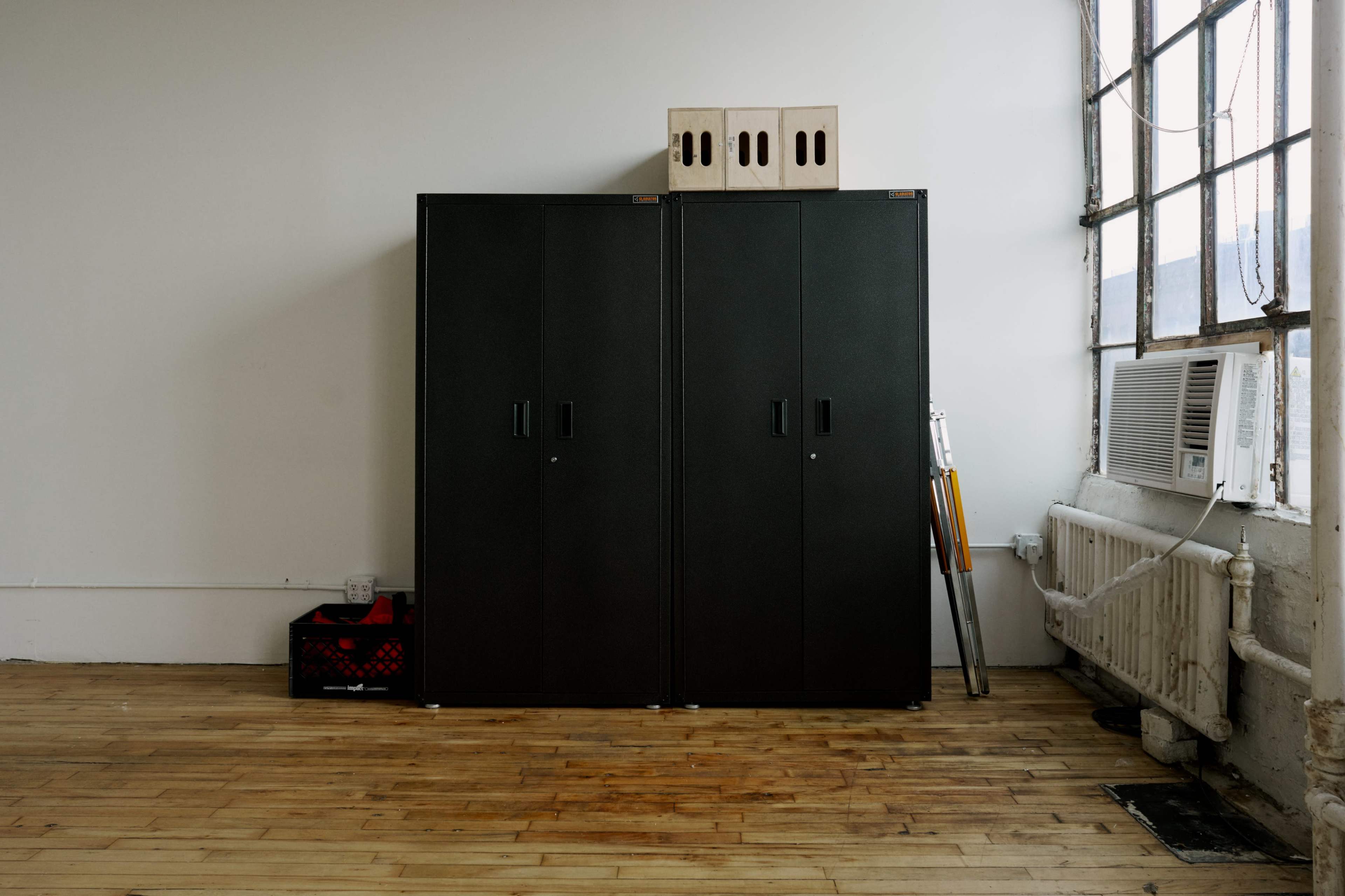 A pair of black cabinets stands against a wall in a spacious room with wooden floors and a window air conditioning unit nearby.