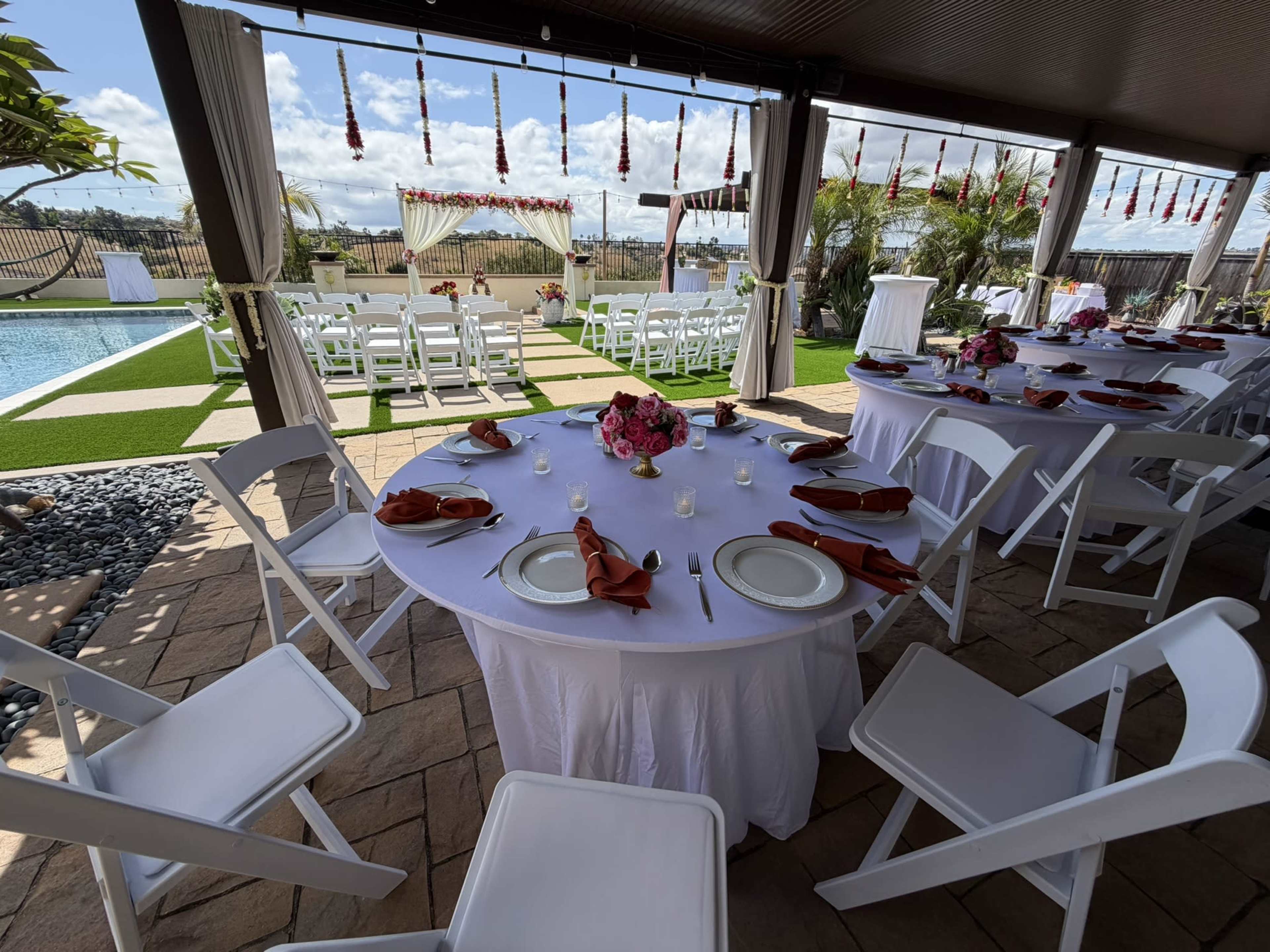The image shows a beautifully arranged outdoor event space with tables set for dining, a decorated altar area for a ceremony, and rows of white chairs positioned around a pool.