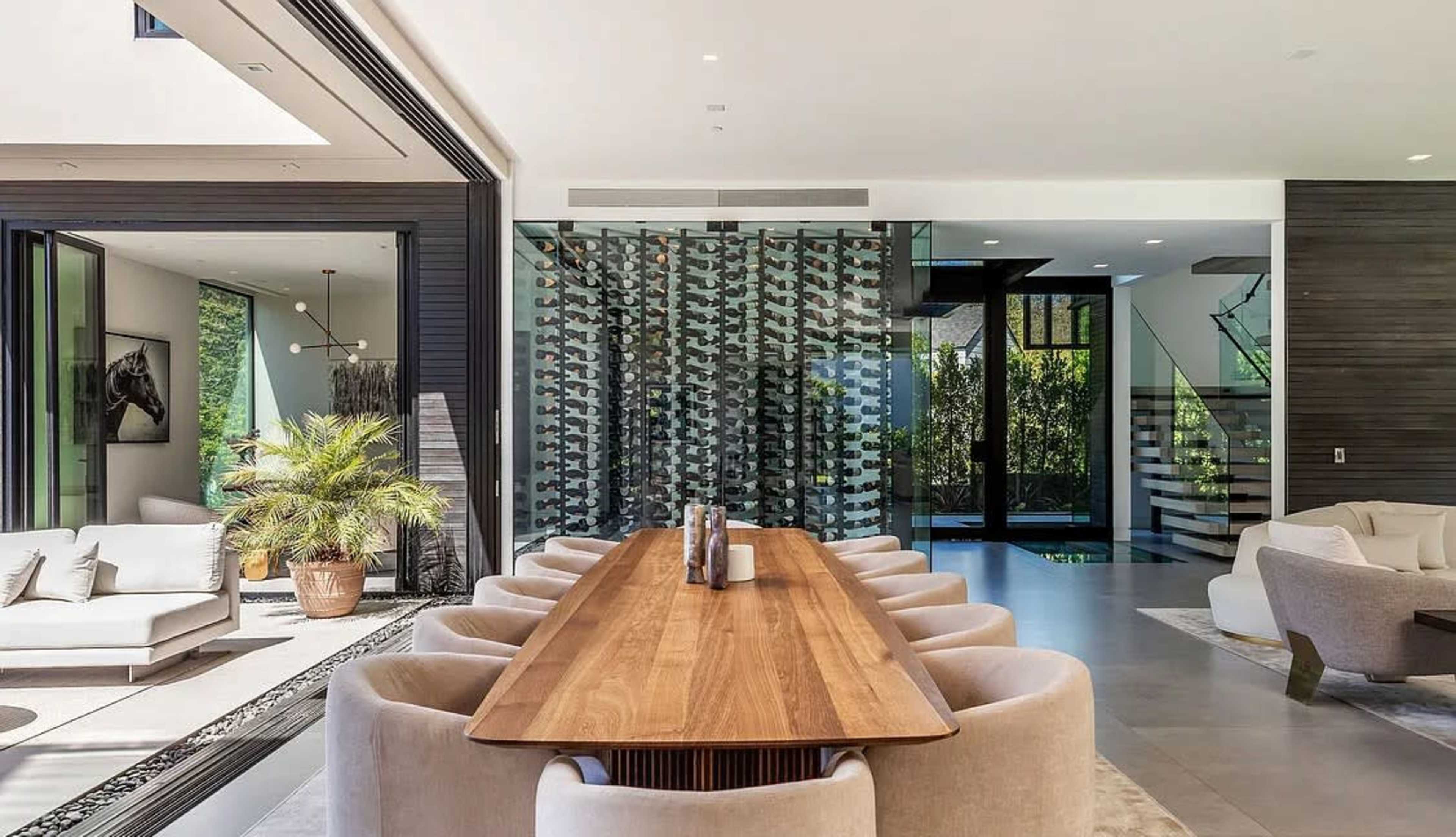 The image shows a modern dining area featuring a long wooden table surrounded by upholstered chairs, with glass walls providing views of an outdoor space and a wine display in the background.