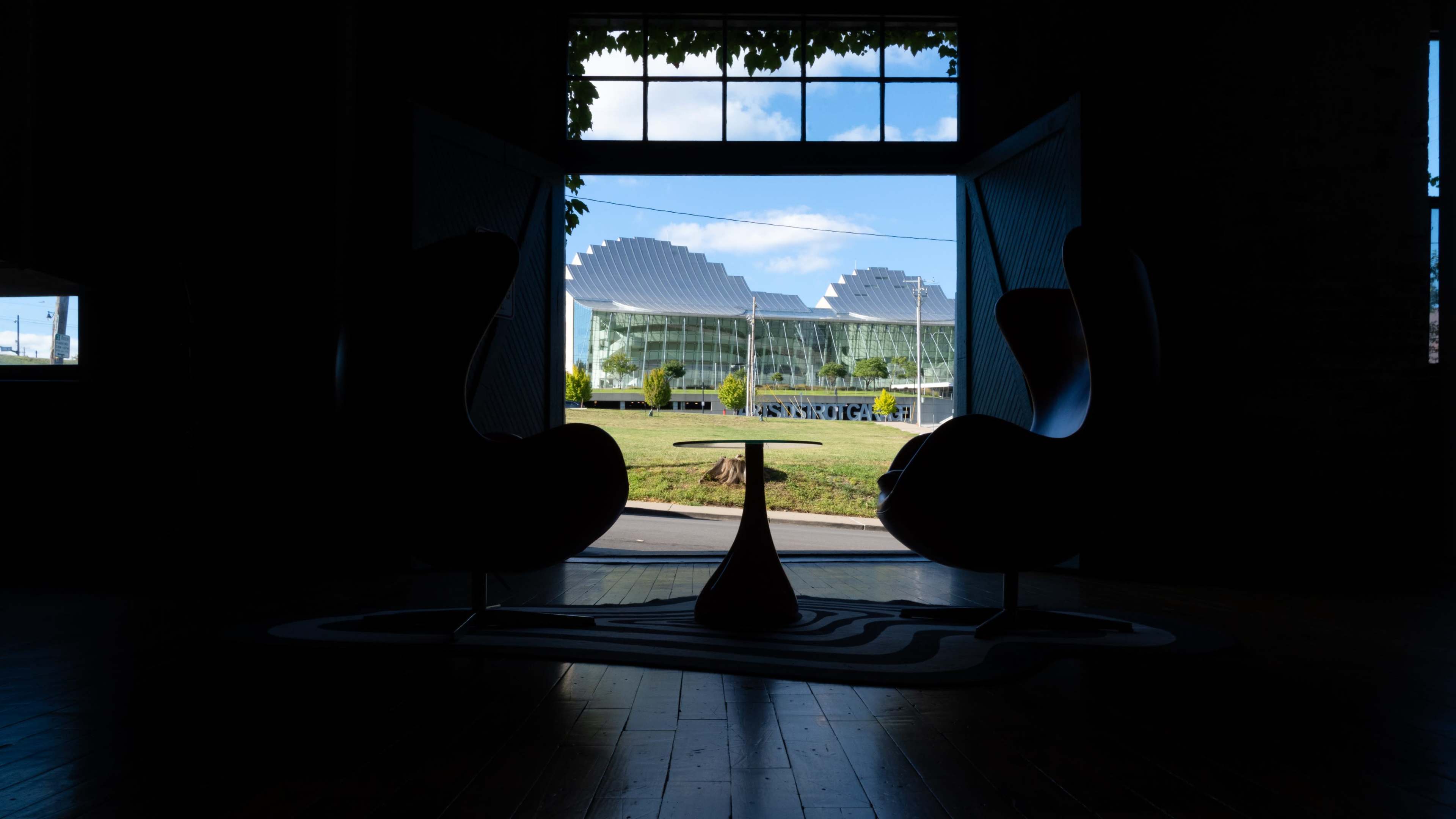 Two chairs are positioned facing a brightly lit doorway, leading to a view of a modern building with a distinctive roof design and a grassy area outside.