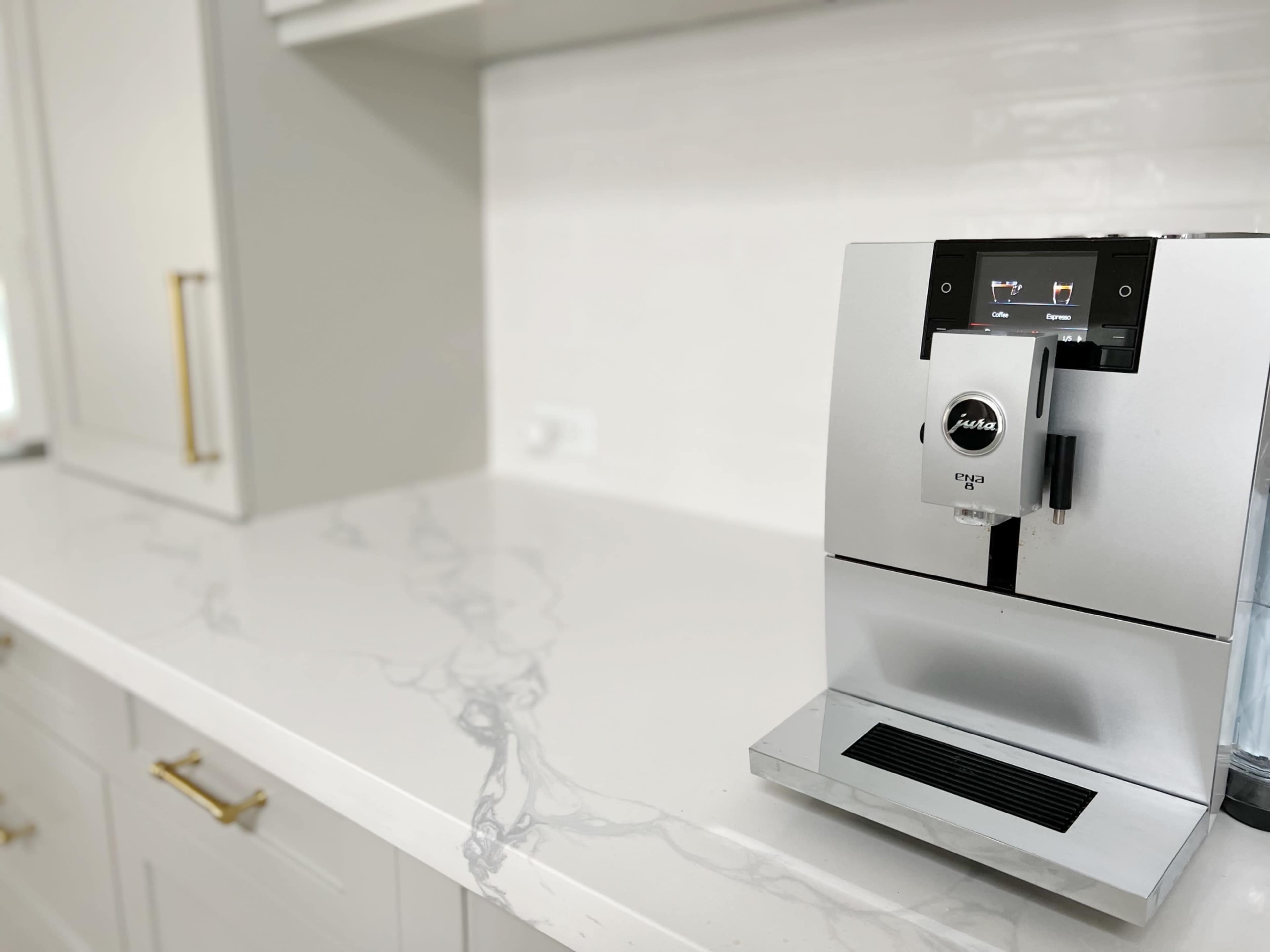A silver coffee machine is positioned on a white countertop with gray veining in a modern kitchen.