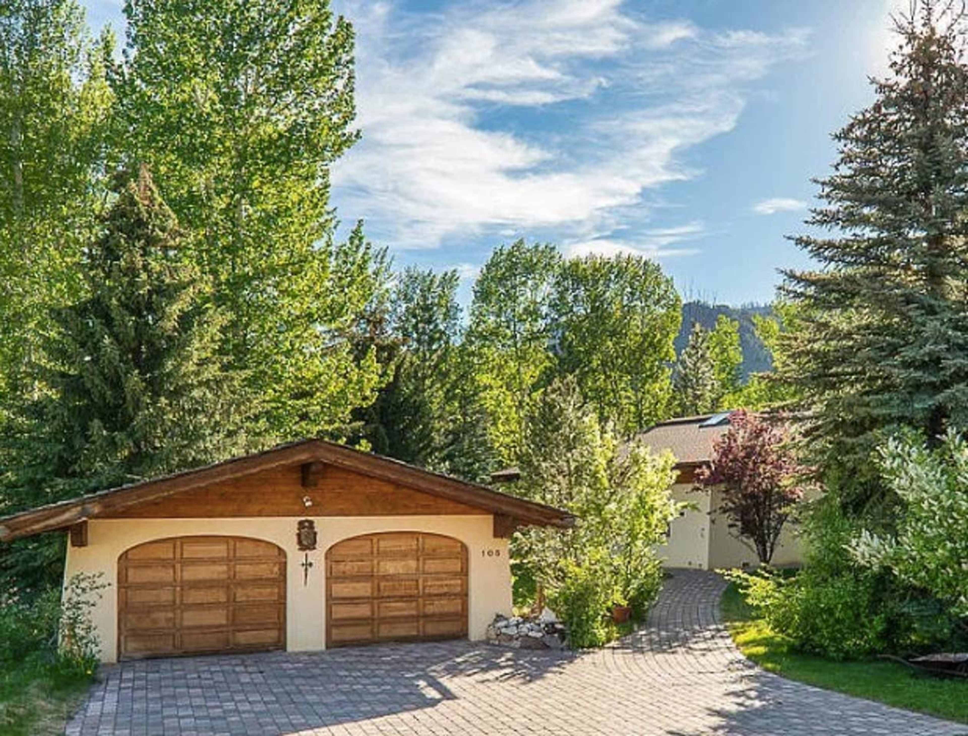 A house with a wooden garage and a paved driveway is surrounded by tall trees and greenery under a blue sky.