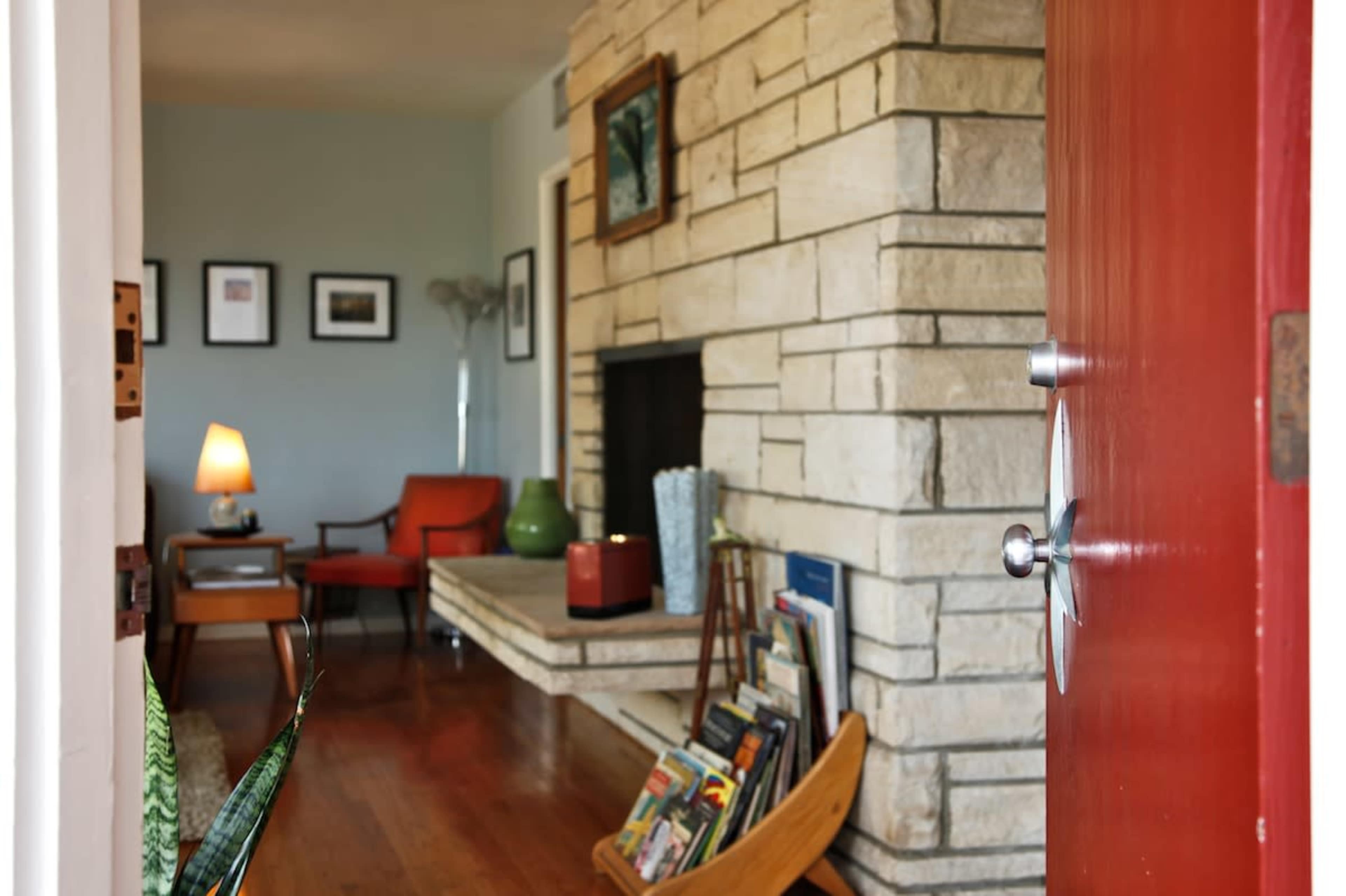 The image shows an interior hallway leading to a living room with a stone fireplace, a red door, and various pieces of furniture.