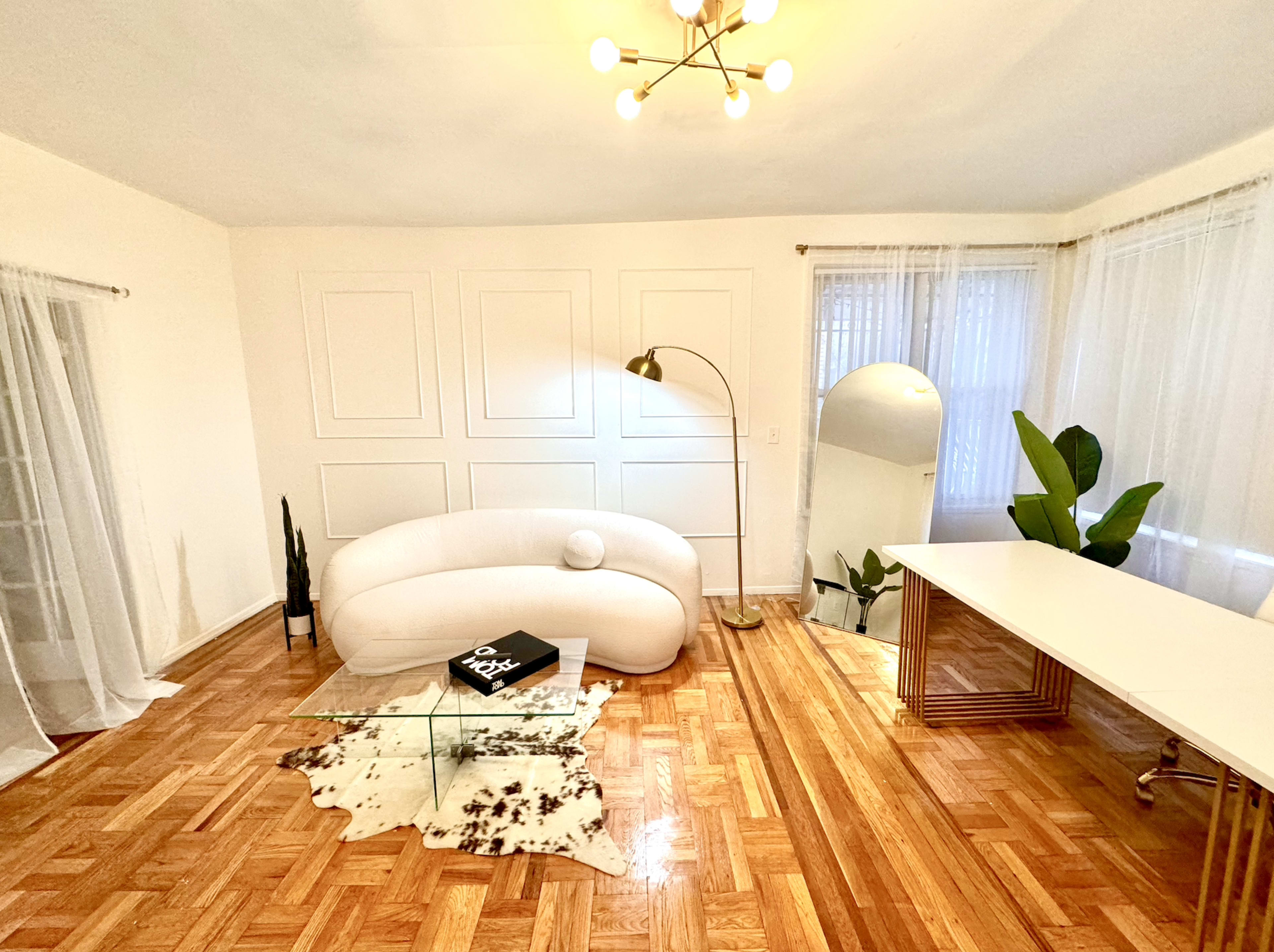A modern, minimalist living space features a curved white sofa, a glass coffee table on a cowhide rug, a workspace with a desk, a large mirror, and potted plants.