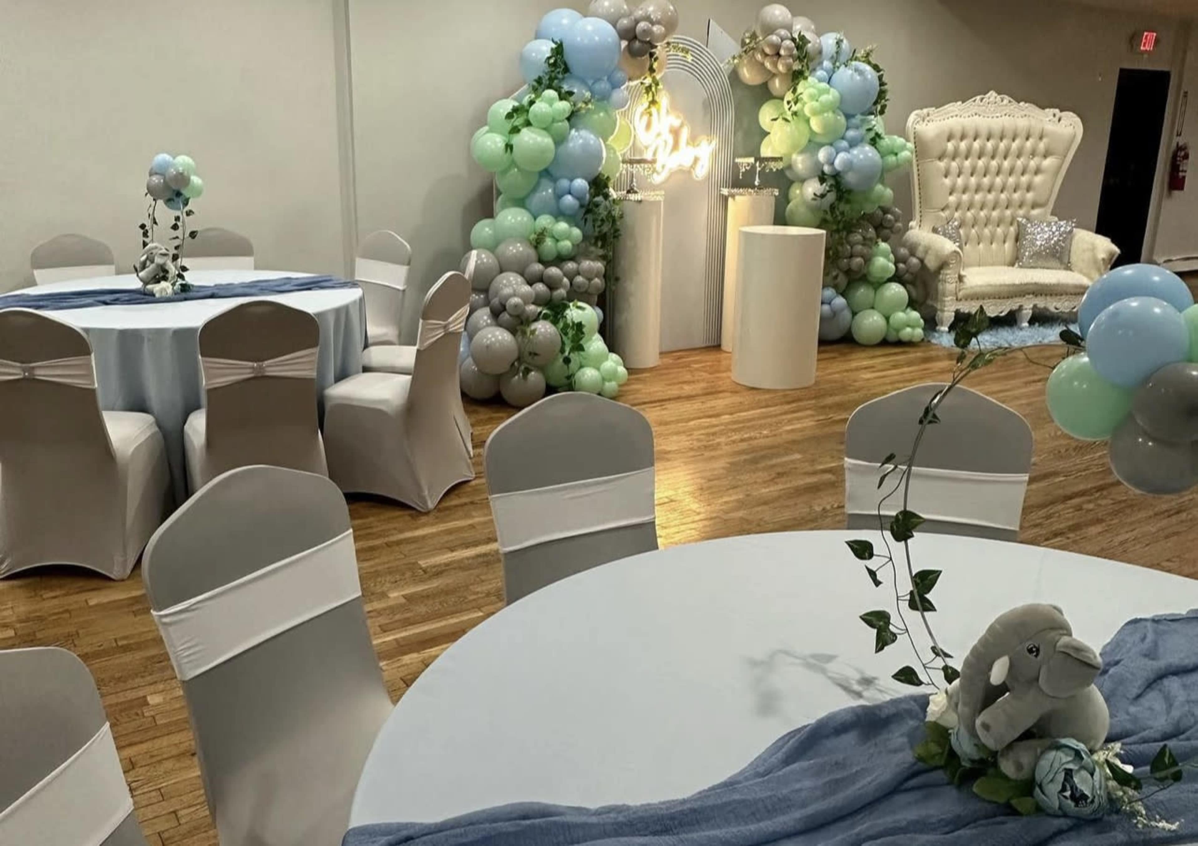 A decorated event space features tables with light blue and white linens, a large balloon arrangement in pastel colors, and a plush white seating area.