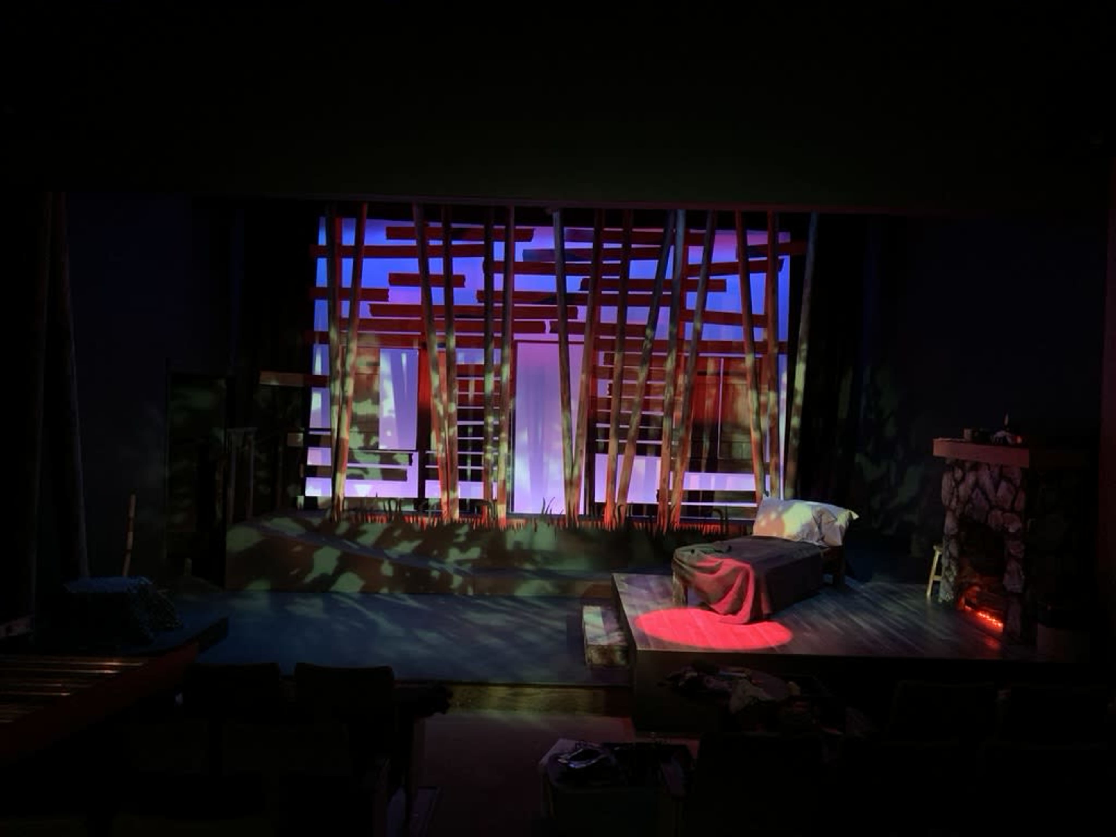 A theater stage set with a bed, a stone fireplace, and dramatic lighting effects creating a backdrop of vertical structures that resemble trees.