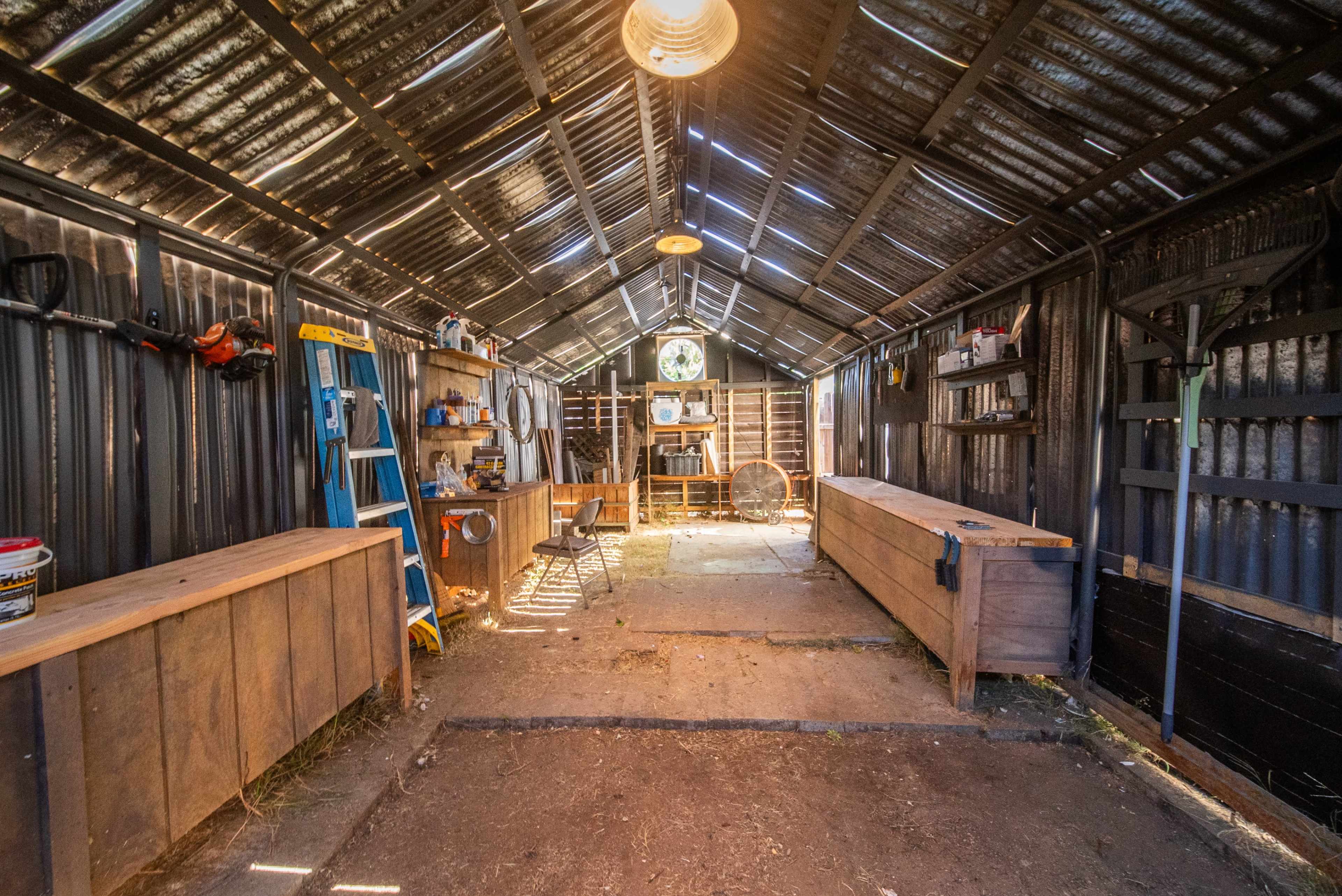 The interior of a metal shed features wooden workbenches, shelves with tools, and a ladder against the side walls.