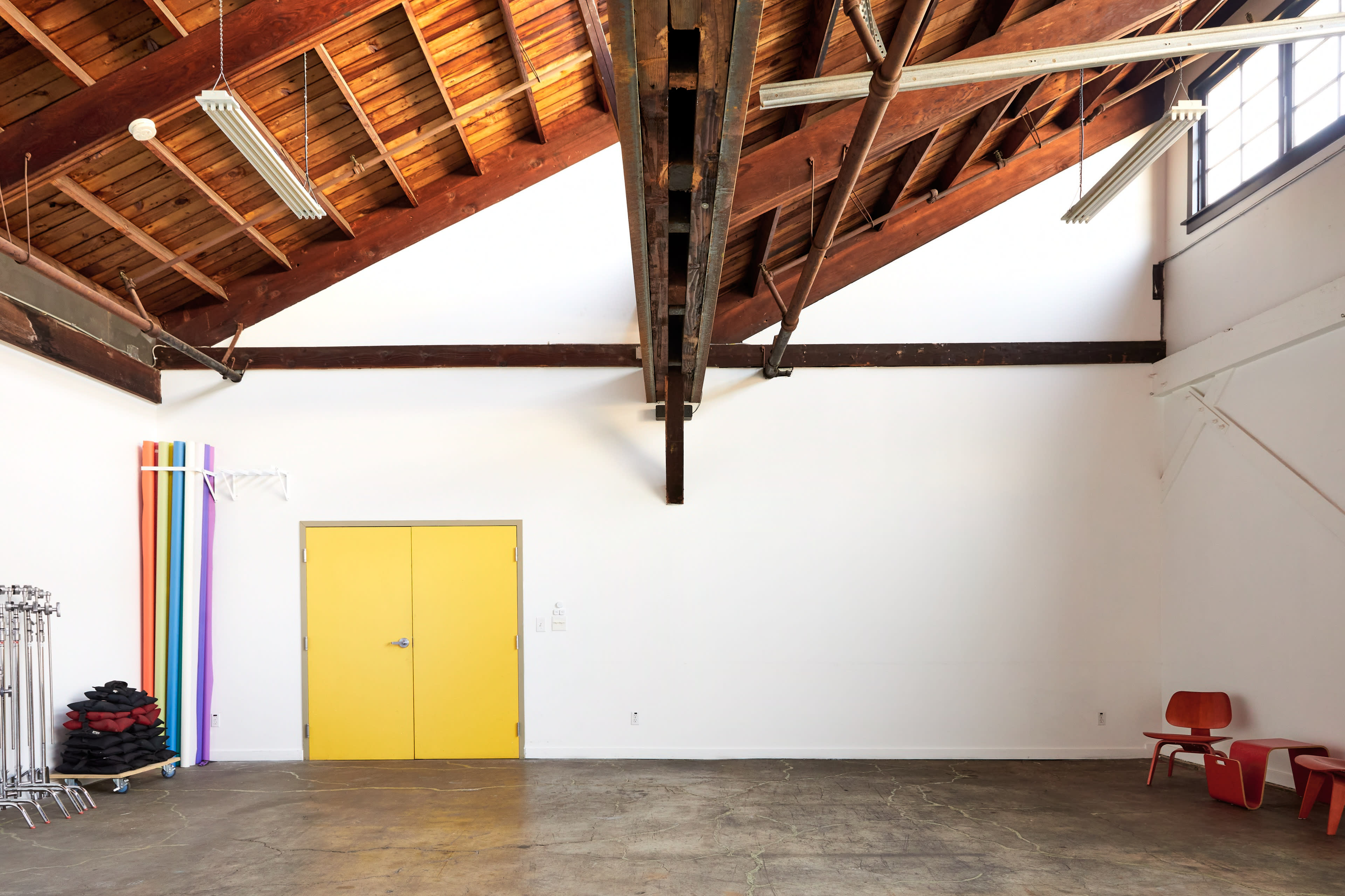 A spacious room with a high wooden ceiling, featuring a yellow door, colorful vertical fabric drapes, and a few red chairs arranged against a white wall.