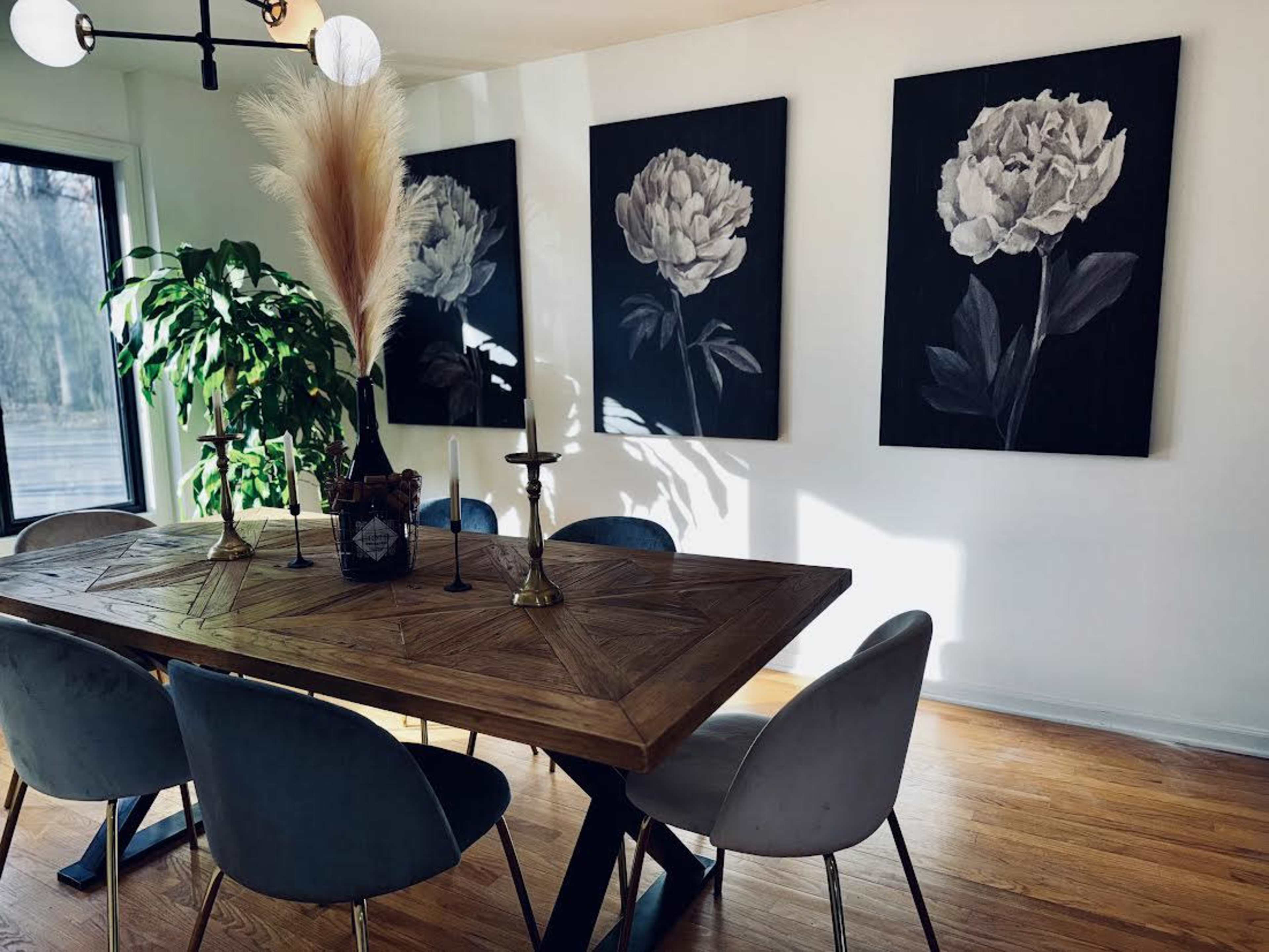 A wooden dining table with candles and decor is surrounded by upholstered chairs in a well-lit room featuring large flower paintings on the walls.