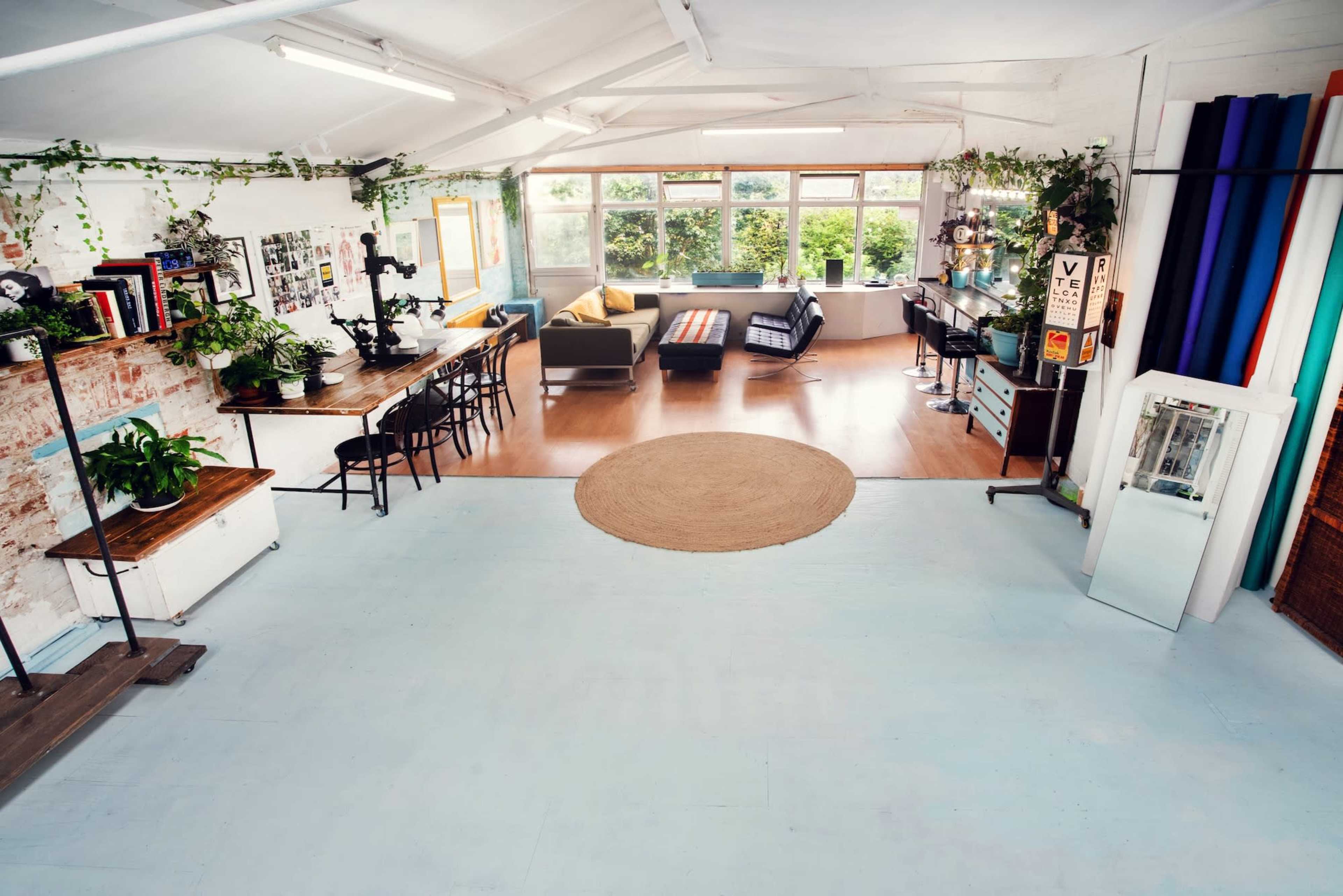 A spacious, well-lit studio with a mix of plants, a long dining table, seating areas, and large windows.
