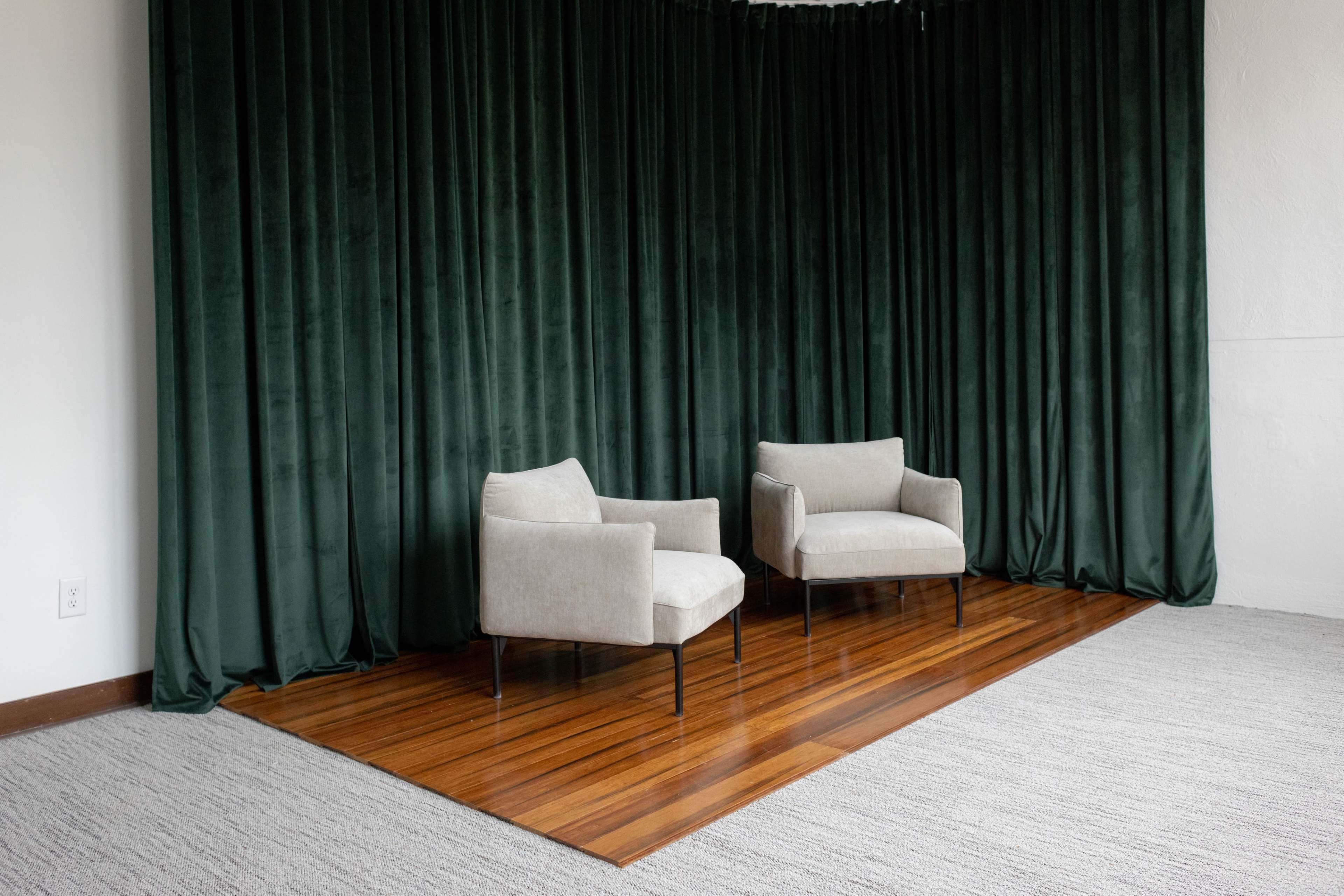 The image shows two beige chairs positioned on a polished wooden platform in front of dark green curtains.