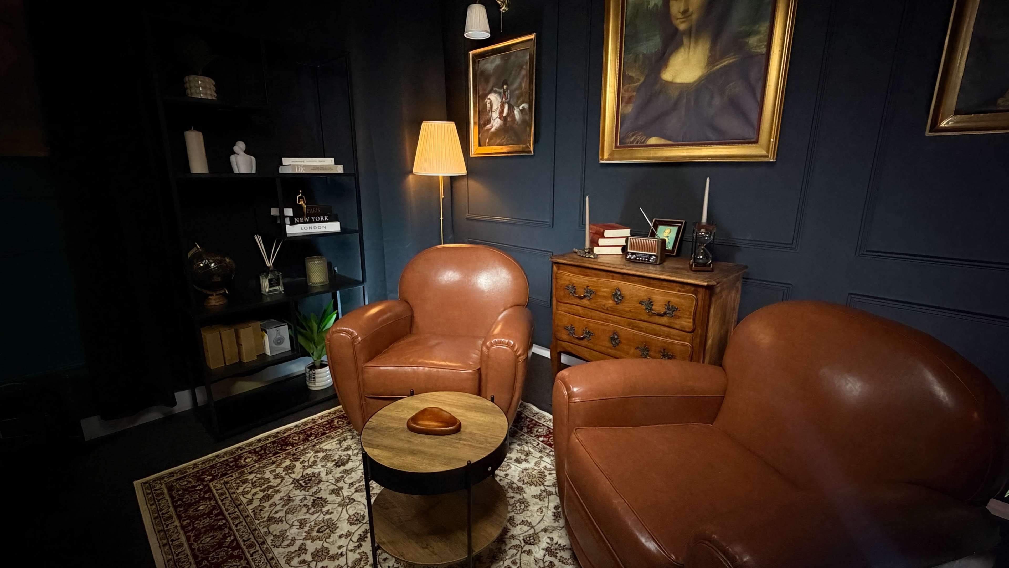 The image shows a cozy corner of a room featuring two brown leather armchairs, a small round table, a floor lamp, and a bookshelf against a dark blue wall adorned with framed artwork.