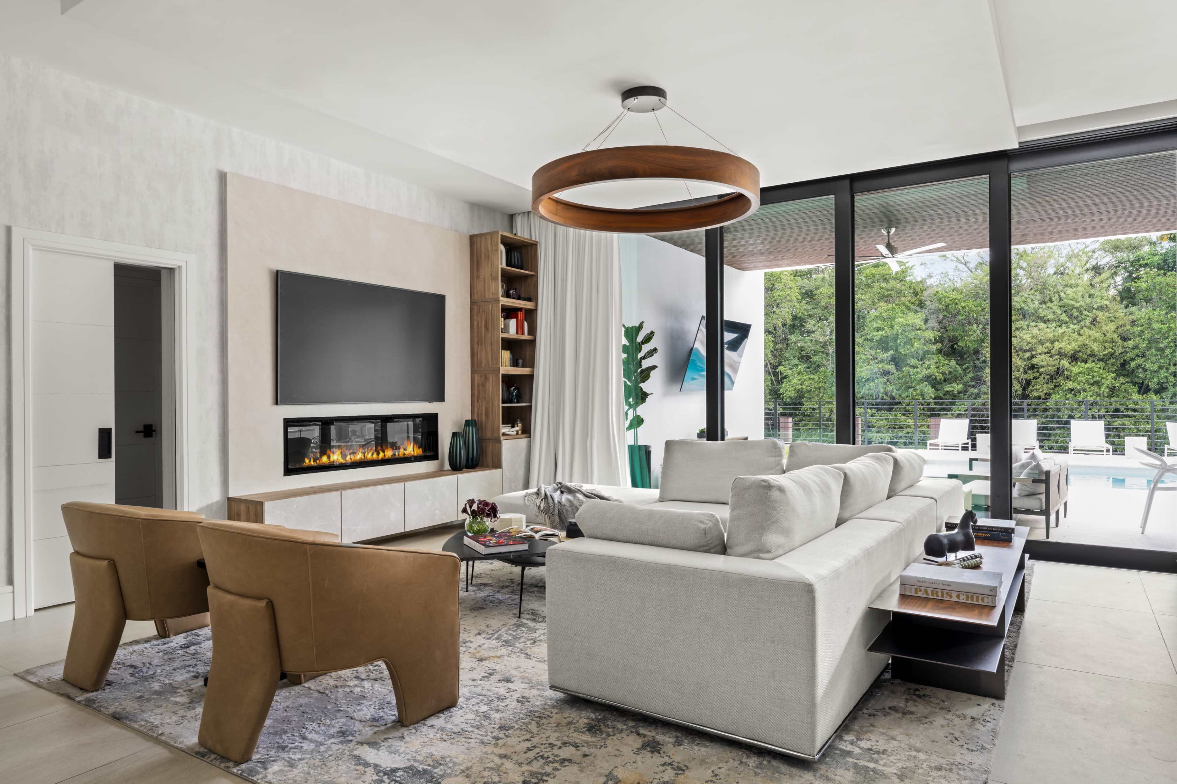 A modern living room features a sectional sofa, two armchairs, a wall-mounted television, and large glass windows overlooking a pool area.