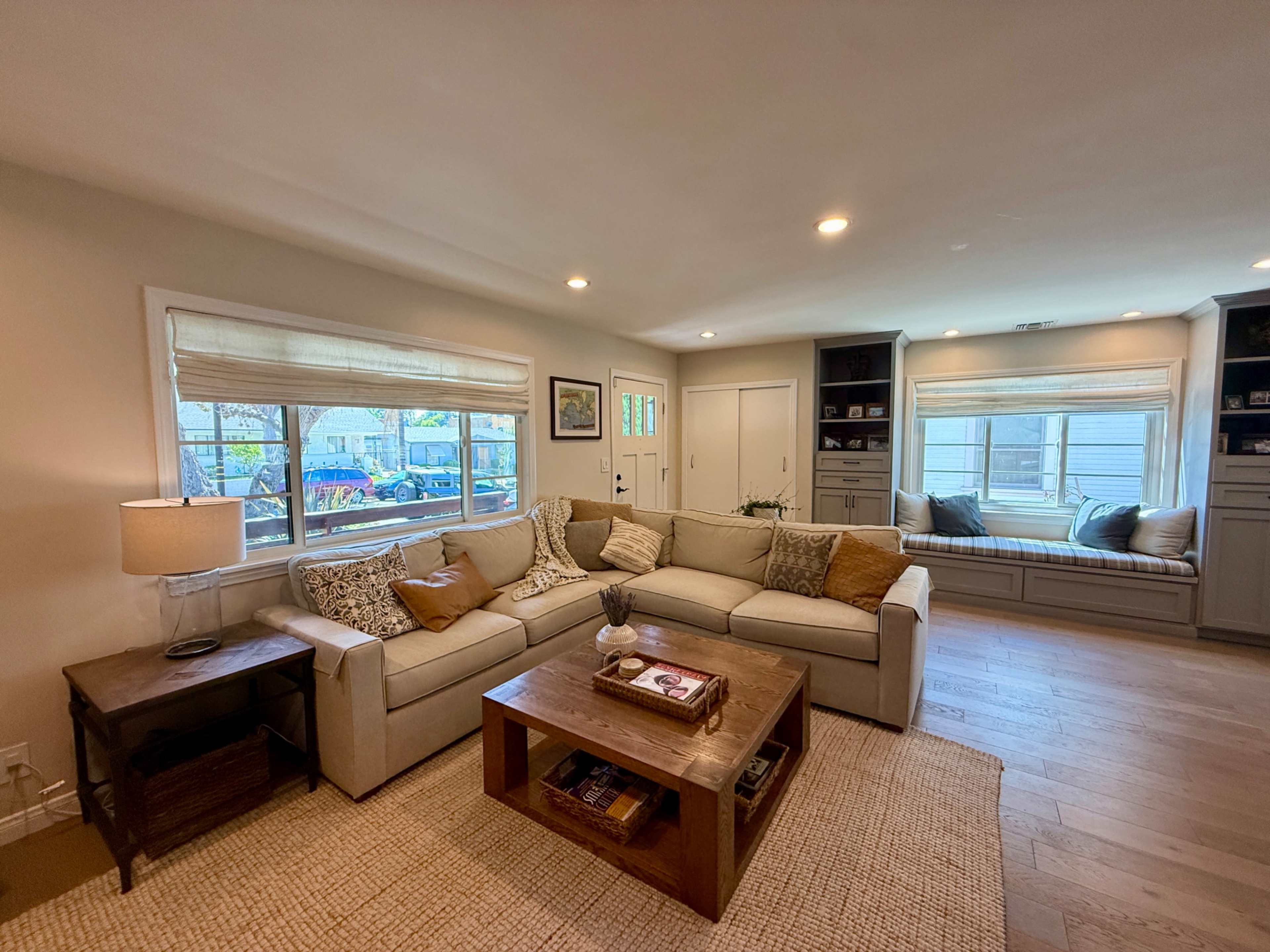 The image shows a cozy living room with a sectional sofa, a wooden coffee table, and a window seat, all well-lit by natural light from large windows.
