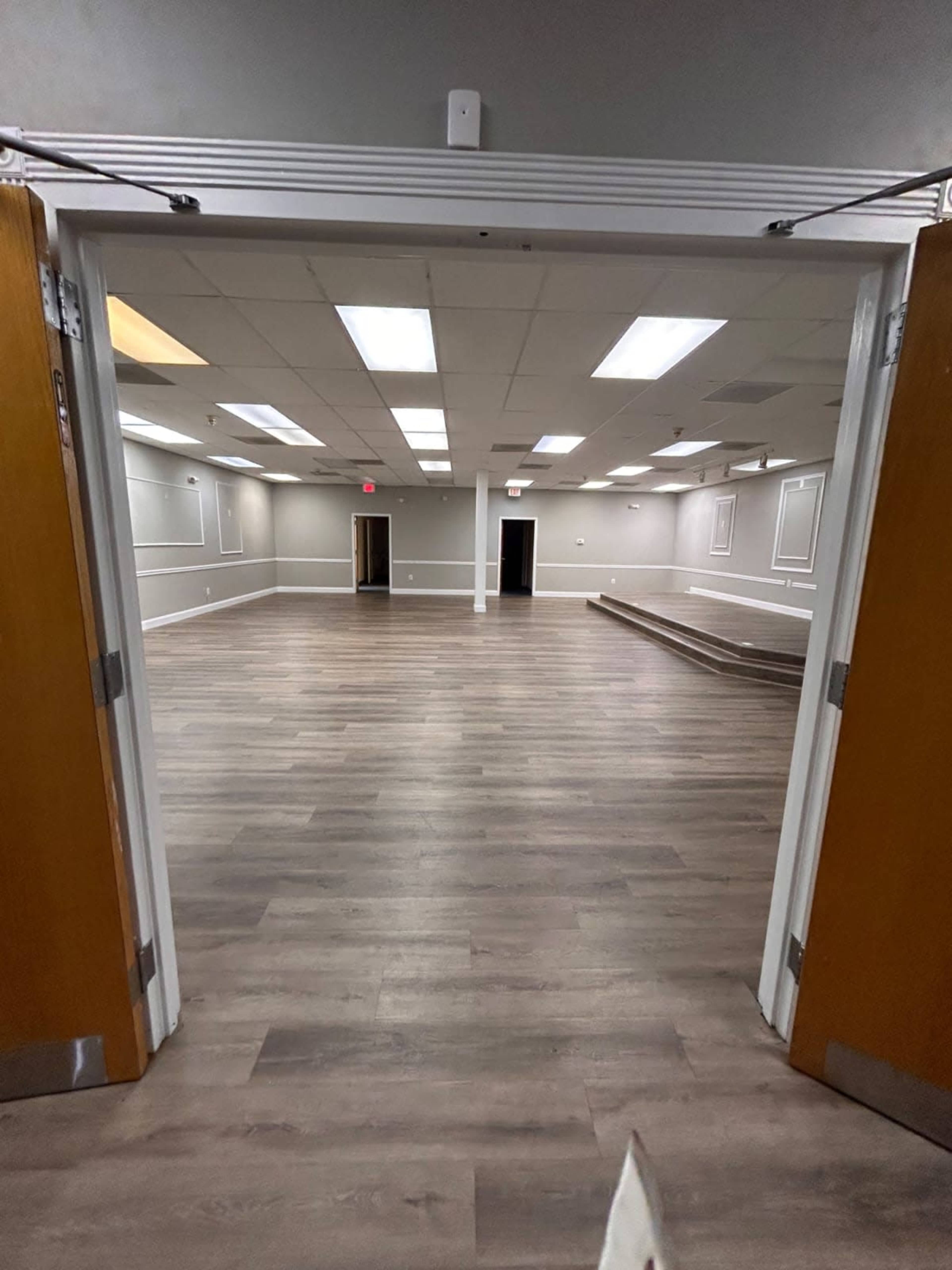 The image shows a large, empty room with wooden flooring and bright overhead lights, viewed through two open double doors.
