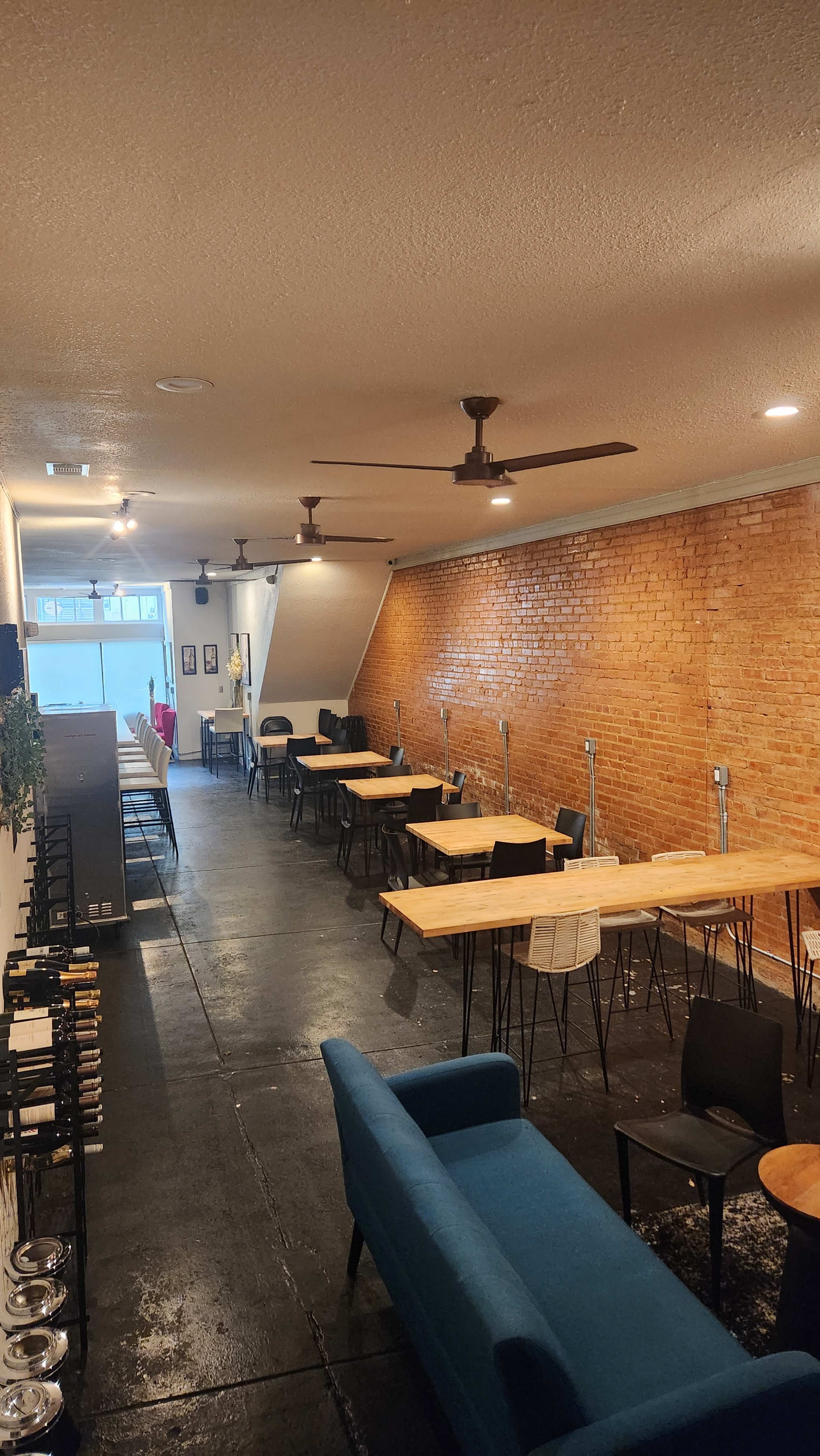 The interior of a narrow café with brick walls, wooden tables, and black chairs arranged along a long aisle.