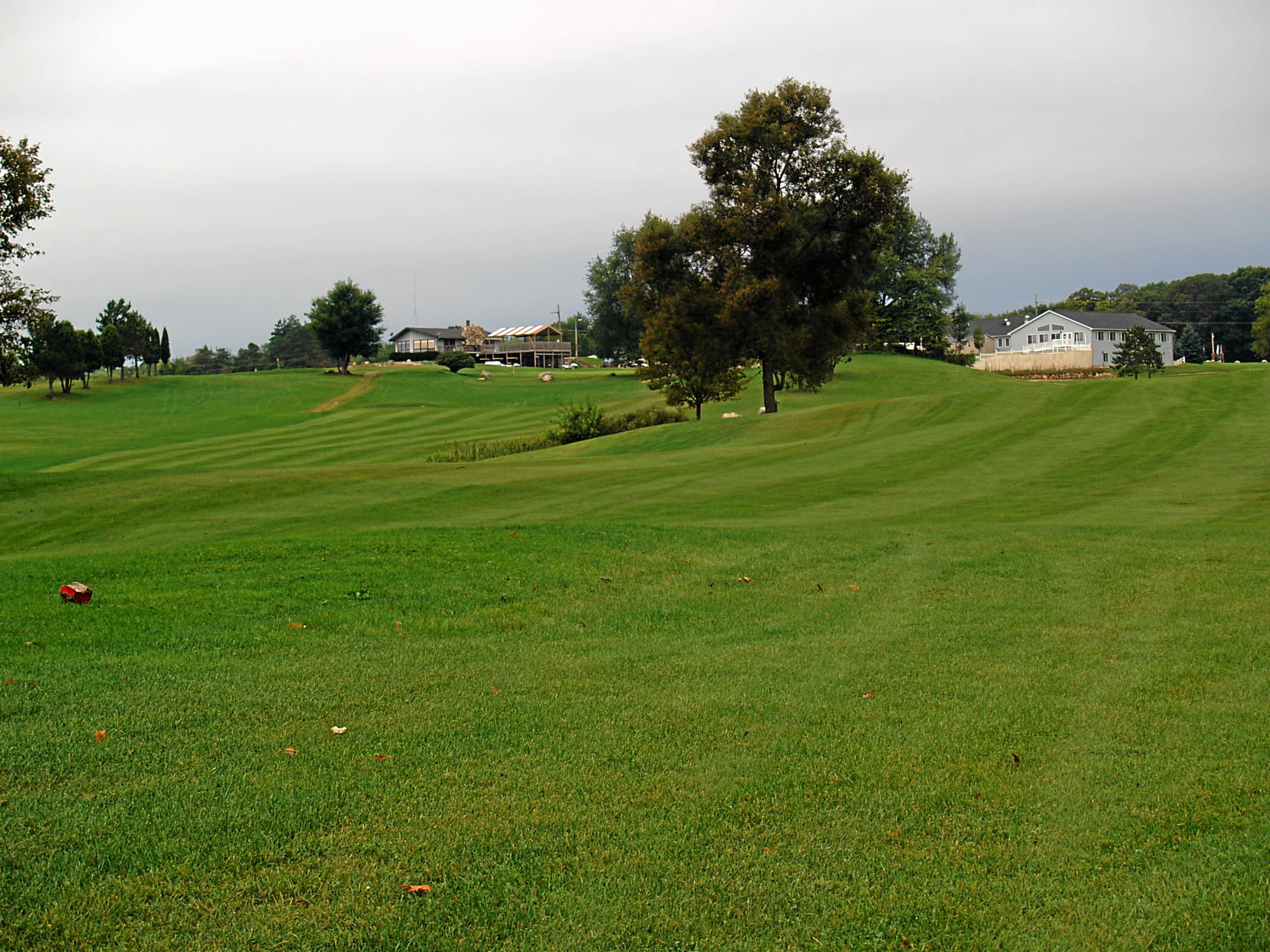 A grassy golf course features rolling hills and a clubhouse in the background, under a cloudy sky.