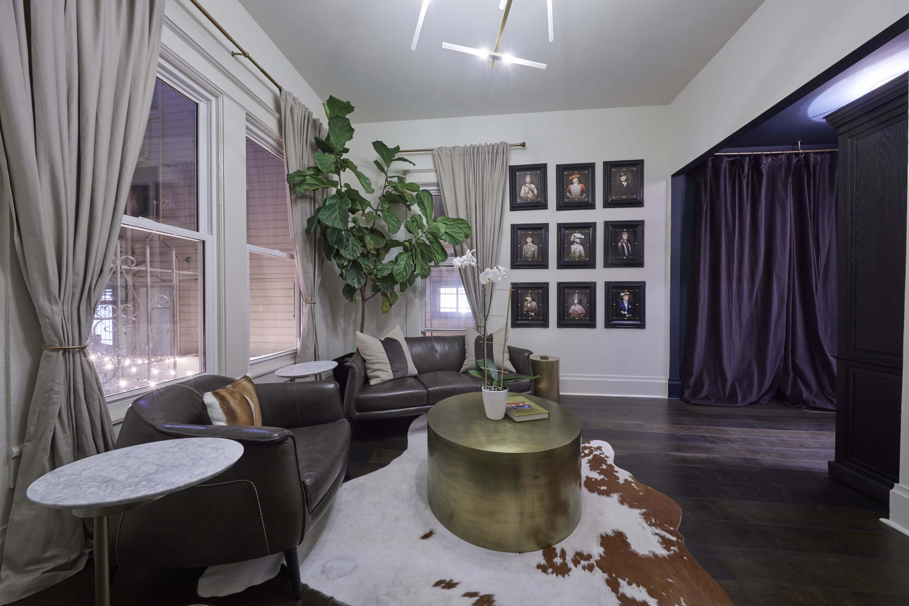 A stylish living room with a circular gold coffee table, a dark leather couch, large windows with sheer curtains, and framed artwork on the wall.
