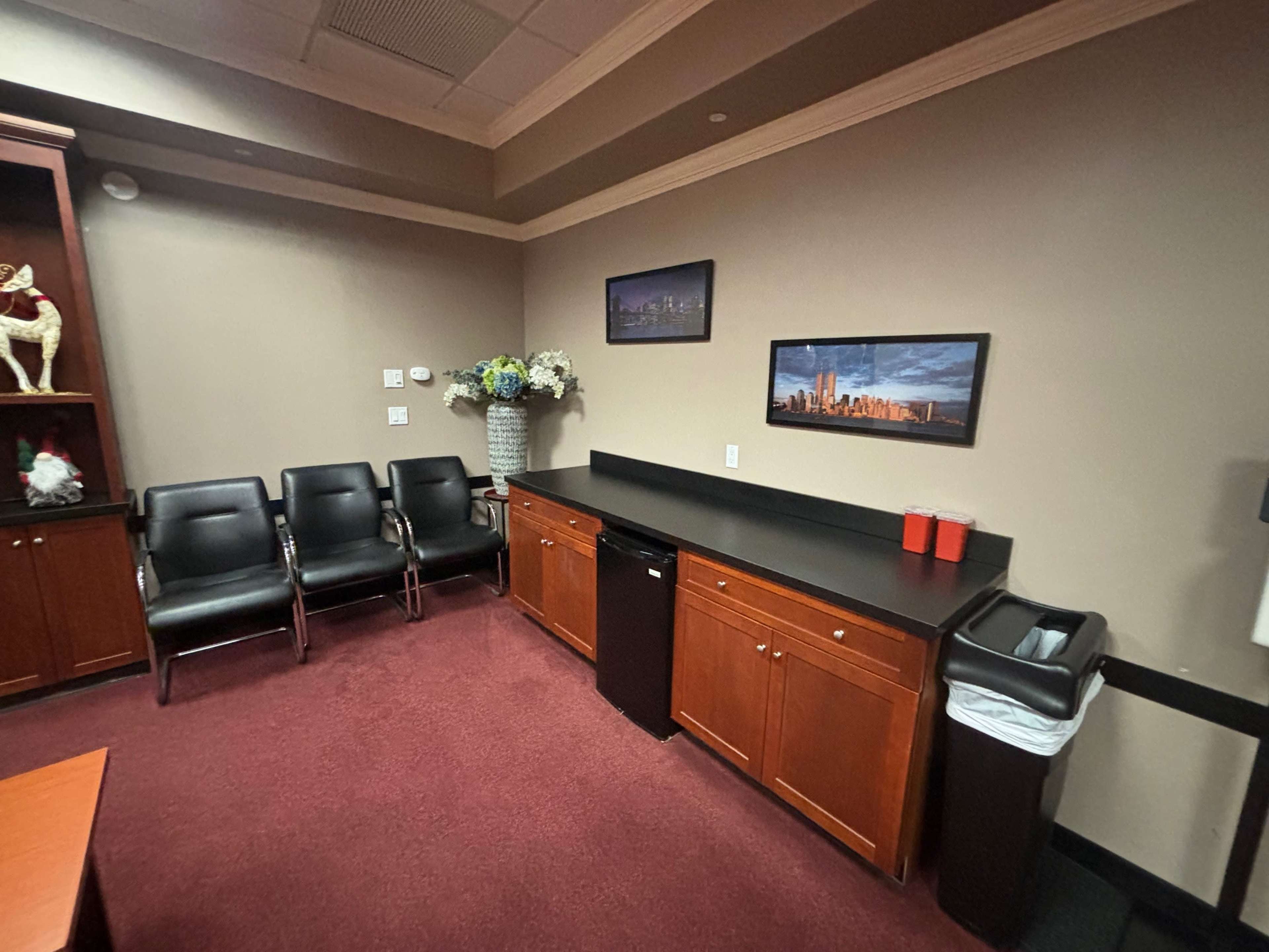 A small office waiting area with three black chairs, a countertop with two red cups, a small fridge, and a waste bin against a beige wall.